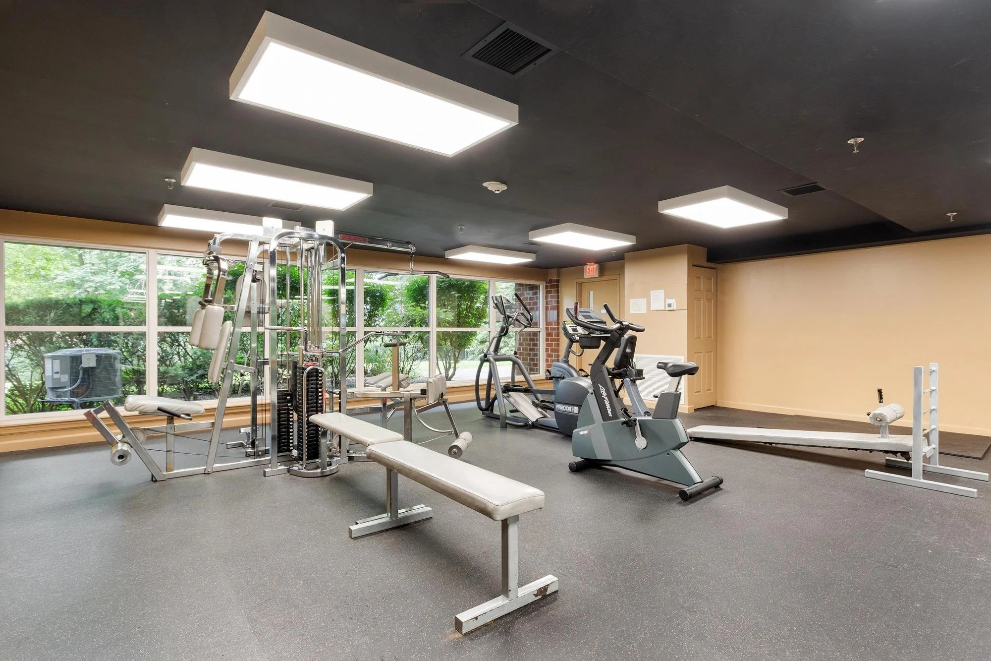 A gym with various exercise machines including a seated bench press, elliptical trainer, stationary bike, and other workout equipment, near large windows with a view of green trees outside.