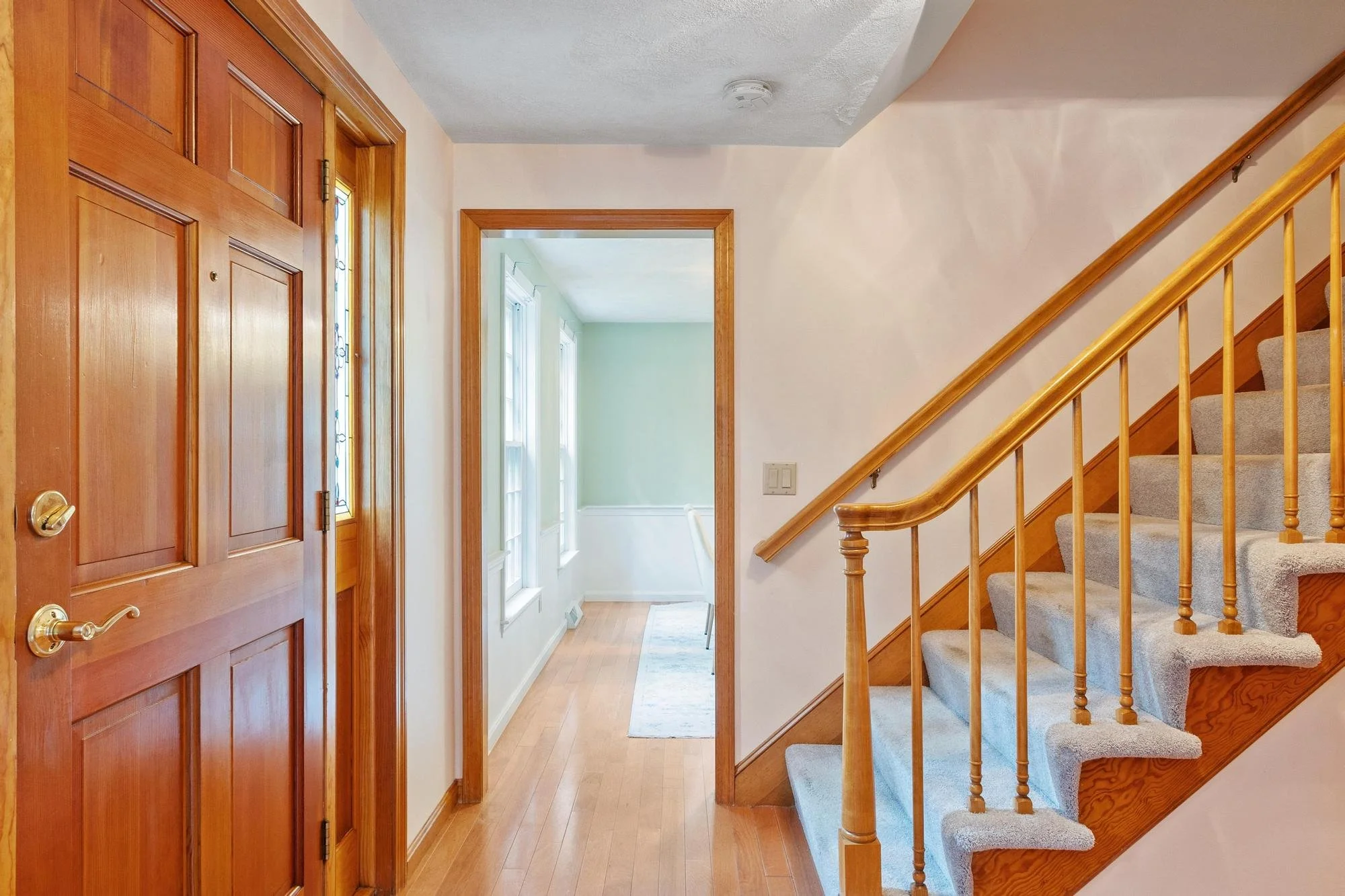 Entryway with a wooden front door on the left, a hallway with windows and natural light at the end, and a staircase with beige carpet and wooden railing on the right.