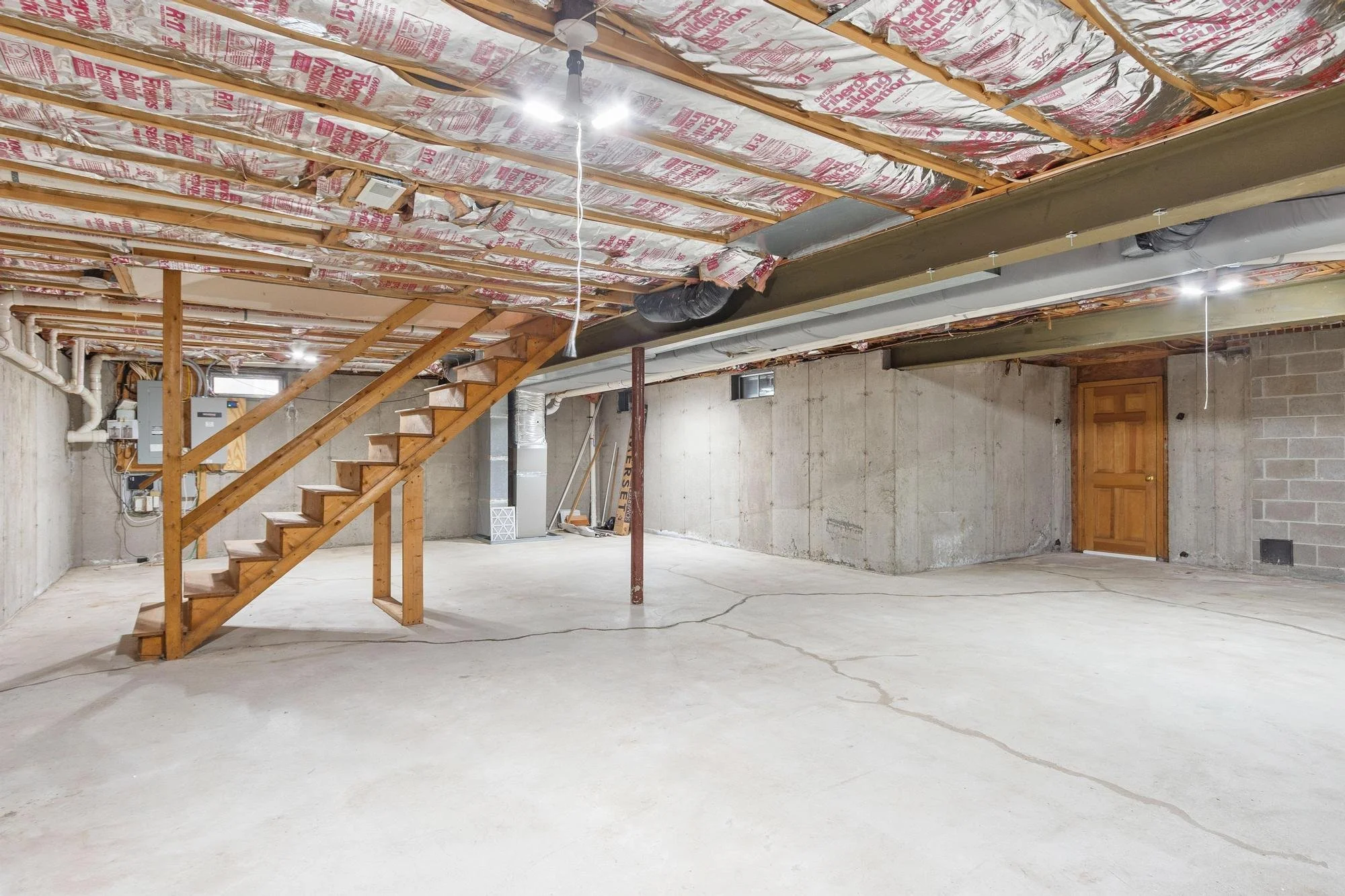 Empty basement with unfinished walls, concrete floor, wooden stairs, and visible insulation in the ceiling.