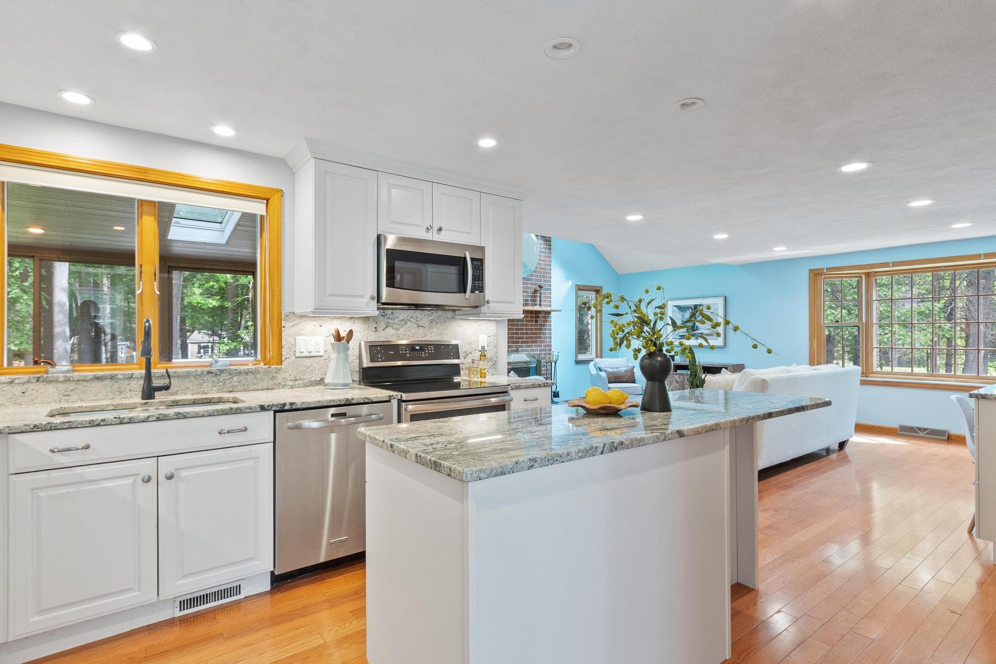 Kitchen with white cabinets and a granite countertop, view into a living room with blue walls and large windows, and a dining area with a vase of yellow flowers.