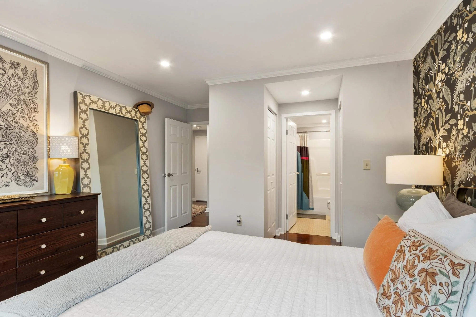 A bedroom with a white bedspread, orange and floral pillows, dark wood dresser, large decorative mirror, lamps, and floral wallpaper on one wall. There's an open closet door and a view into a bathroom with a shower curtain.