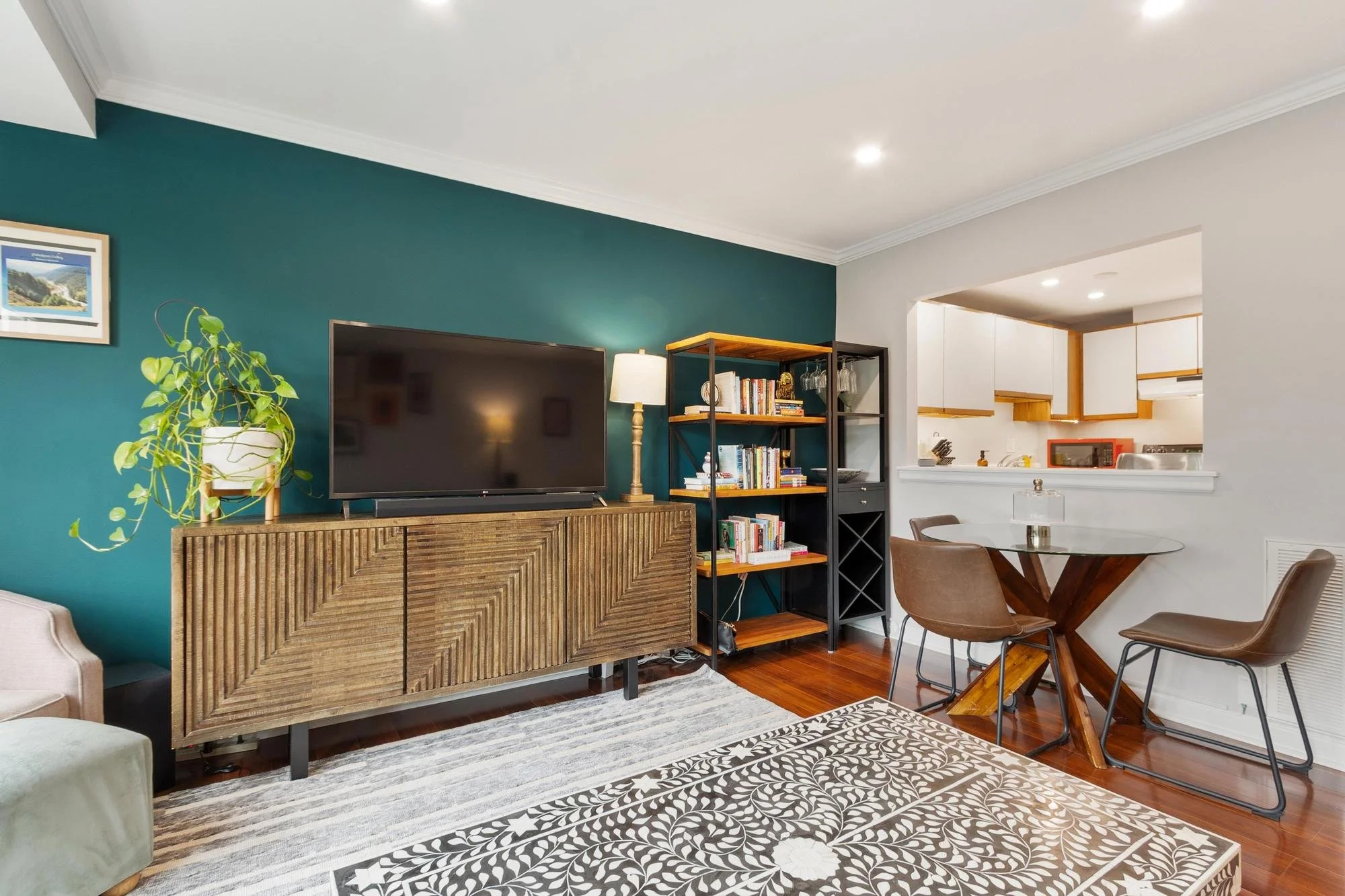 Living room with teal accent wall, wooden console, flat-screen TV, bookshelf, dining table with beige chairs, patterned rug, and an open kitchen in background.