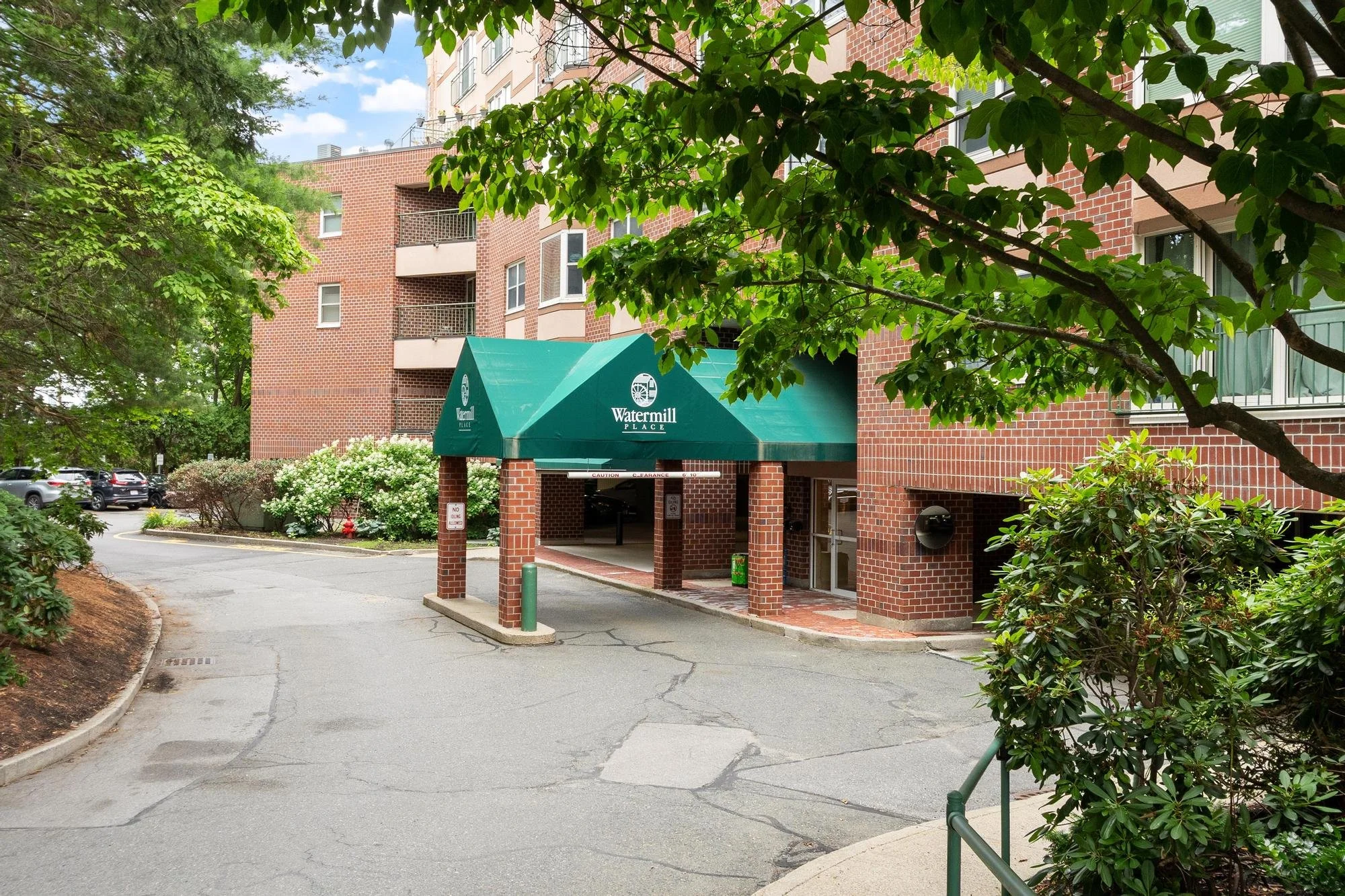 Entrance to Watermill Place residential building with a green canopy over the driveway, red brick walls, and multiple balconies visible in the background. Lush green trees surround the scene.