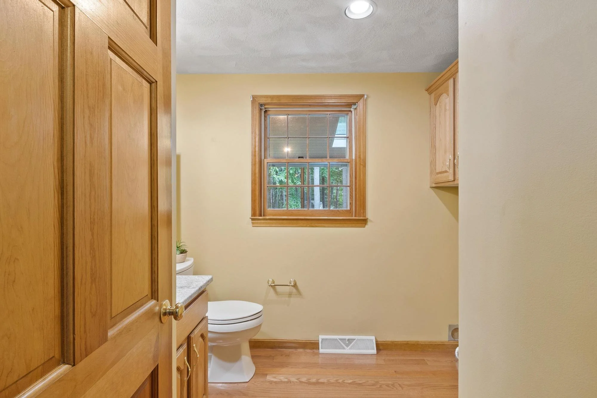 A small bathroom with beige walls, wooden window frame and cabinet, white toilet, a small potted plant on the countertop, and a vent on the hardwood floor near the baseboard.