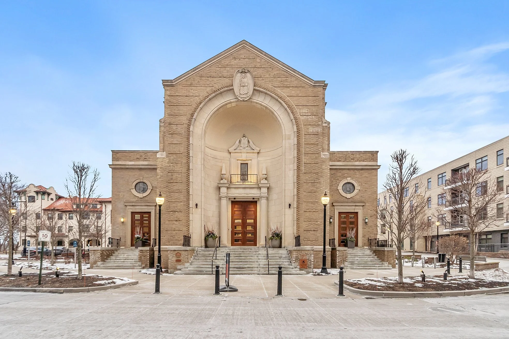 Front view of a church building with stone steps, four lampposts, leafless trees, and residential buildings in the background, on a cold day with snow patches.
