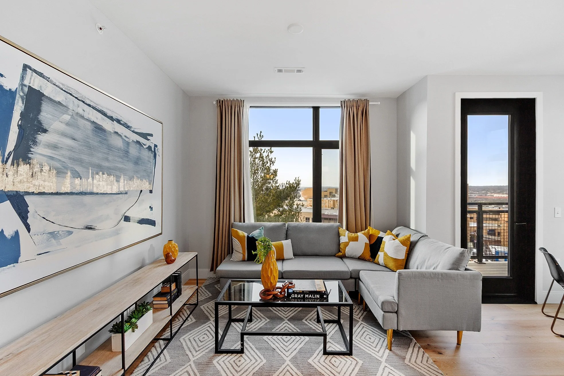 Modern living room with gray sectional sofa, patterned cushions, glass coffee table, abstract wall art, and large windows with beige curtains, overlooking cityscape.
