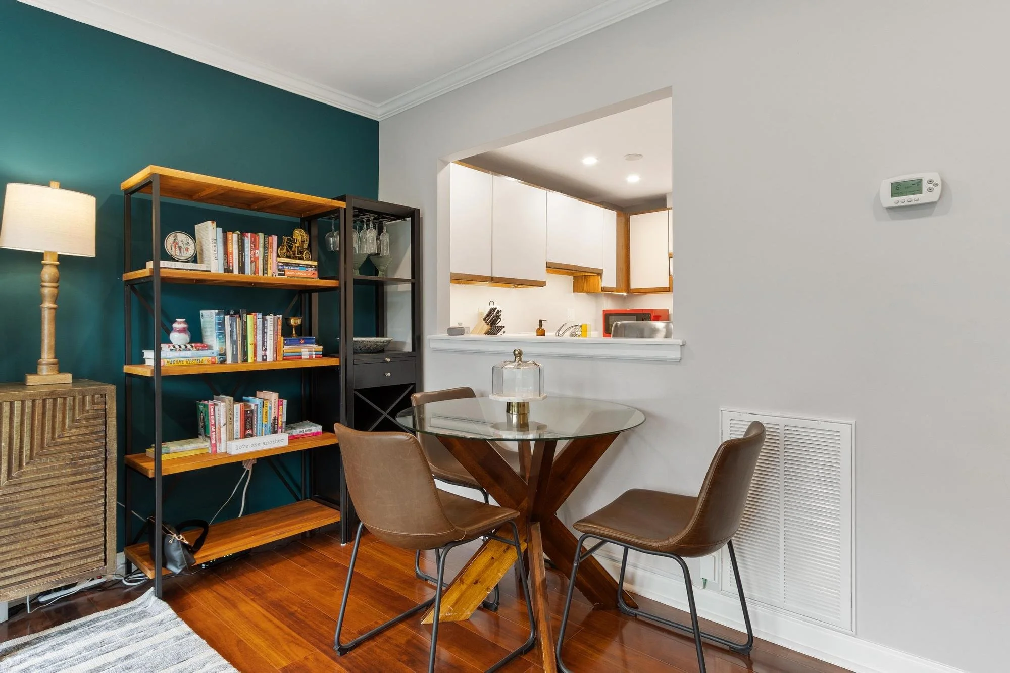 A dining area with a round glass table, four brown chairs, a wooden bookshelf filled with books and decorative items, a lampshade, a wooden cabinet, and a view into a kitchen with white cabinets and a small counter.