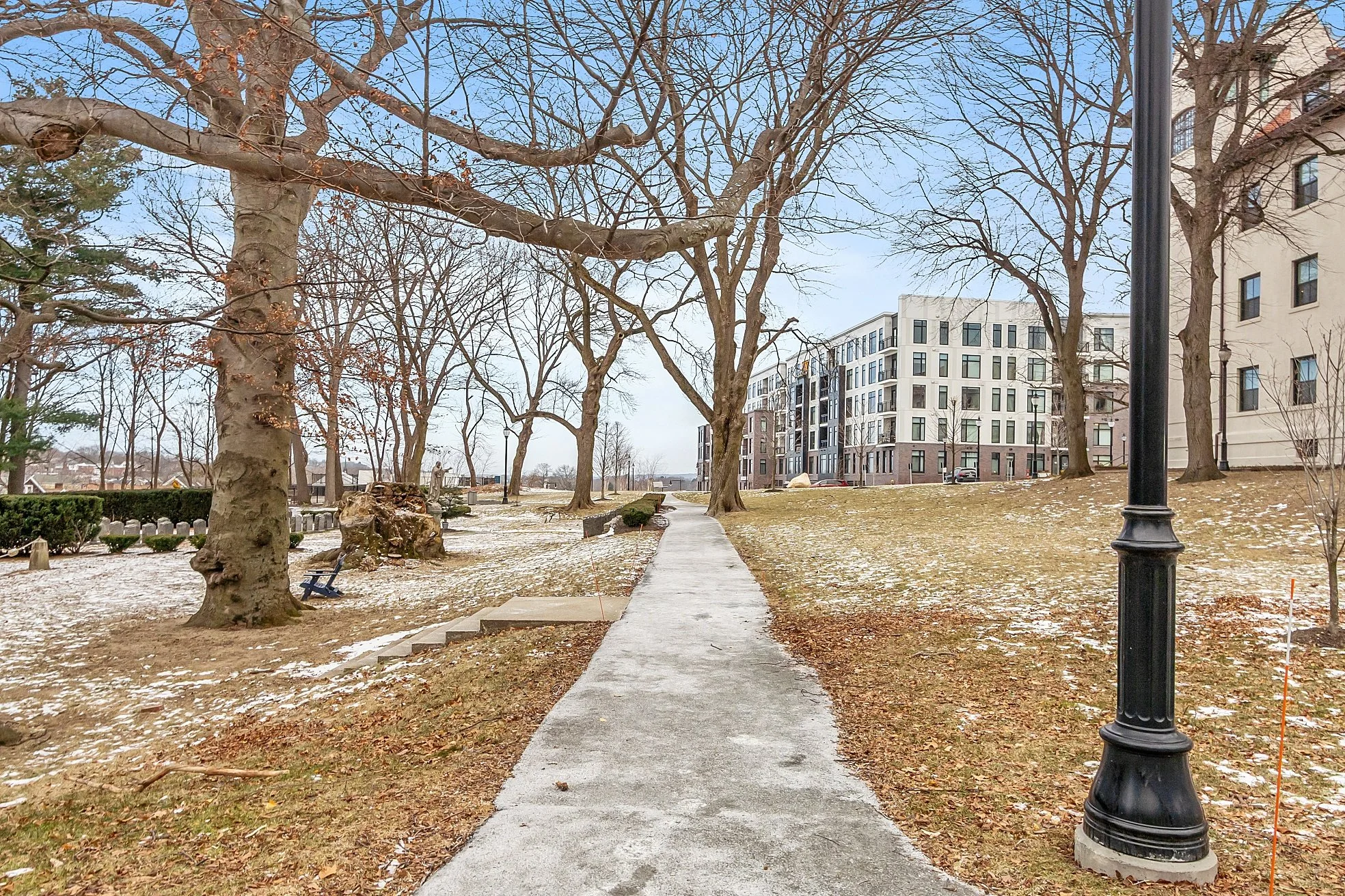 A concrete pathway runs through a park with leafless trees and some patches of snow on the ground. In the background, there are multi-story residential buildings, a black lamppost, and a cloudy sky.
