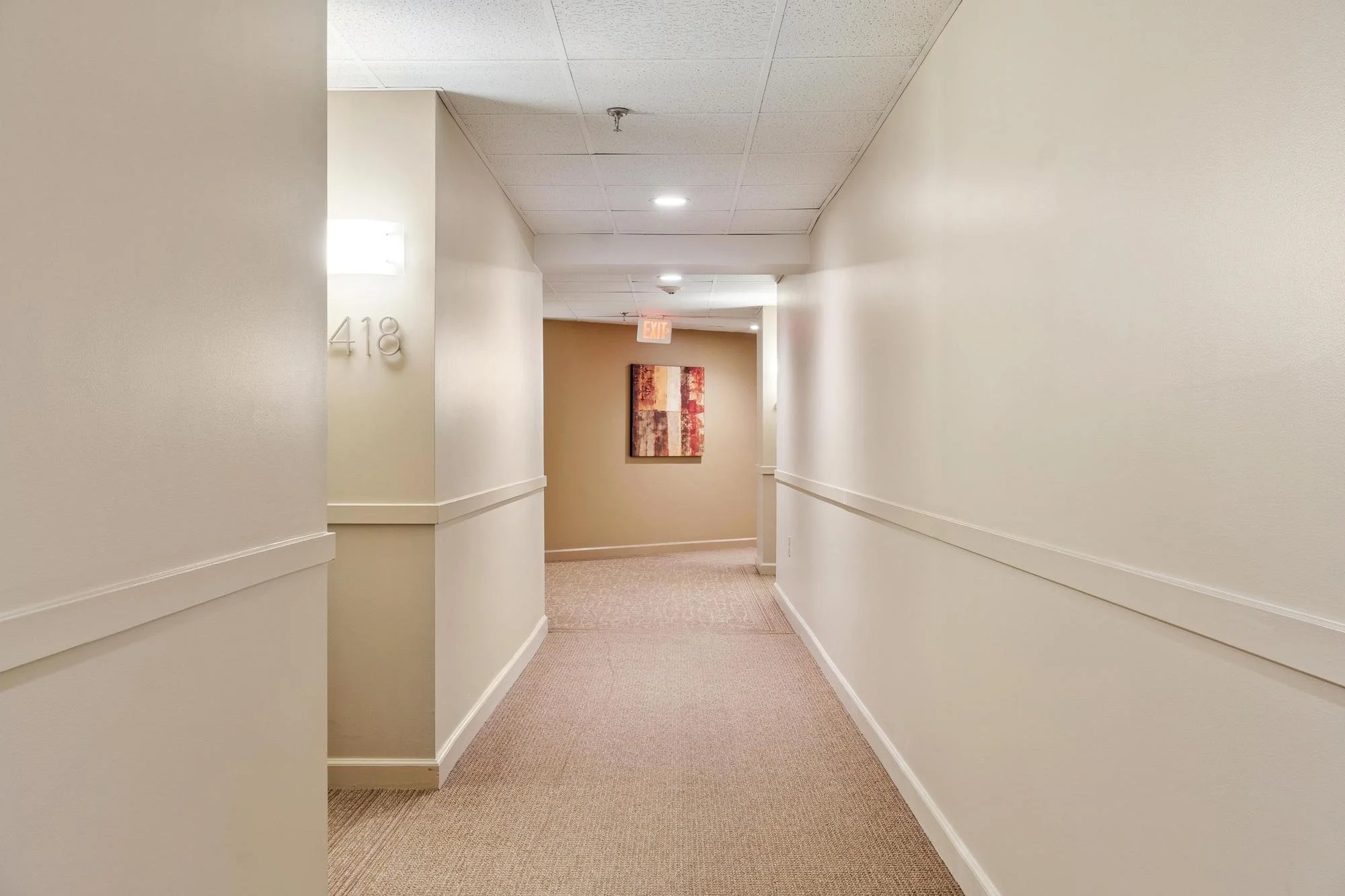 A hallway in an apartment complex or hotel with cream-colored walls, beige carpet, and ceiling tiles. There are wall-mounted lights, a white door with the number 418, and a painting on the far wall. An EXIT sign is visible at the end of the corridor.