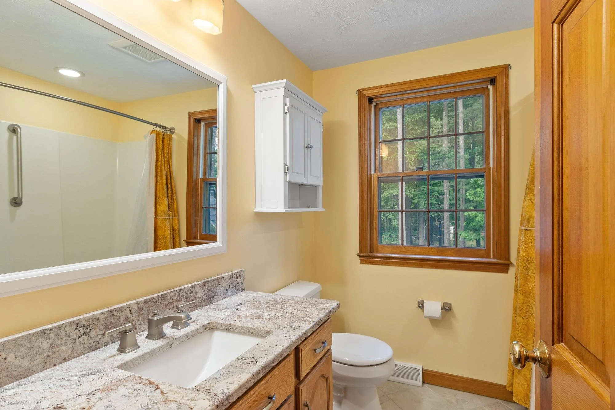 A bathroom featuring a large wall mirror, a granite countertop with dual sinks, a white toilet, wooden cabinetry, a window with view of trees, and yellow walls with yellow curtains.