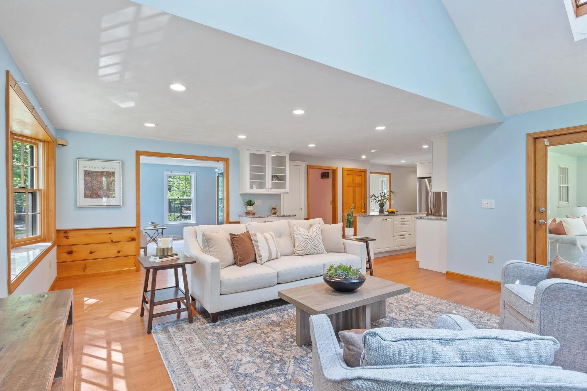 Open-concept living room with light blue walls, white ceiling with recessed lighting, wooden trims, large windows, with a white sofa, light gray armchair, and wooden coffee table on a patterned area rug.