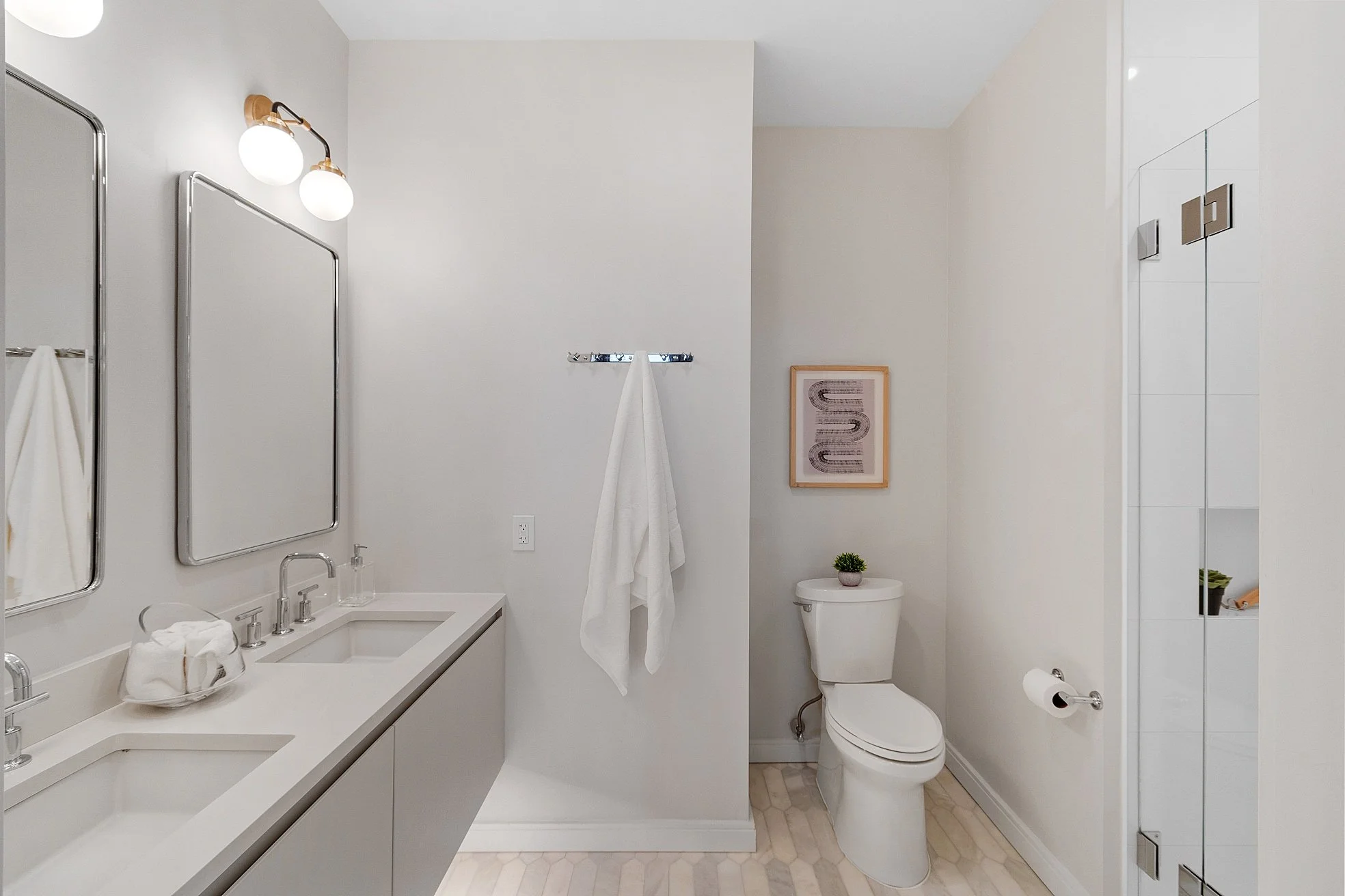 A modern bathroom with a double sink vanity, two mirrors, and two light fixtures above. There is a white towel hanging on a rack, a framed artwork on the wall, and a toilet with a small potted plant on the tank. Part of a shower with a glass door and a built-in shelf is visible.