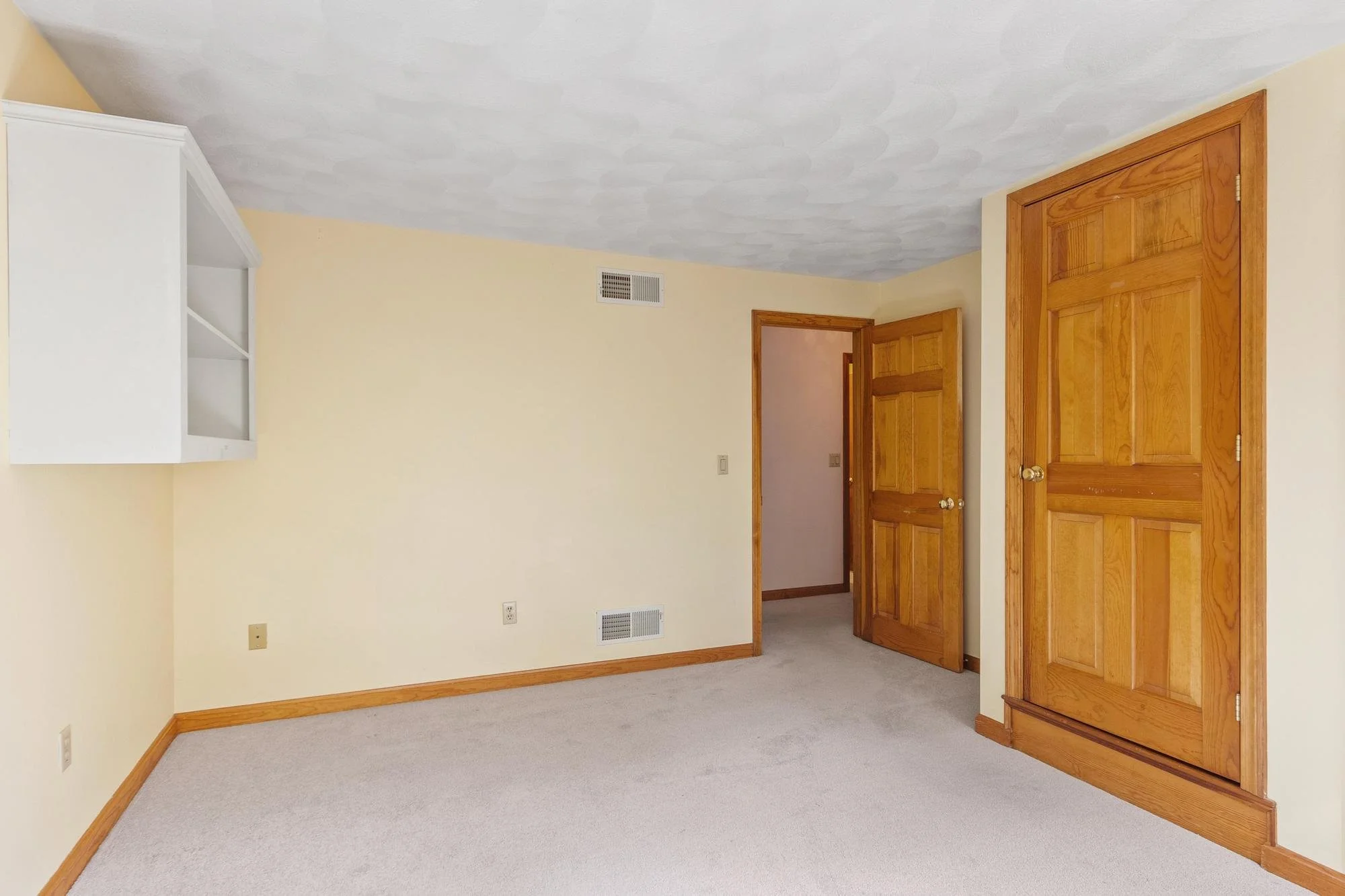 Empty room with beige walls, wooden trim, carpeted floor, and a wooden closet door. There is a small white wall-mounted cabinet and two vents.