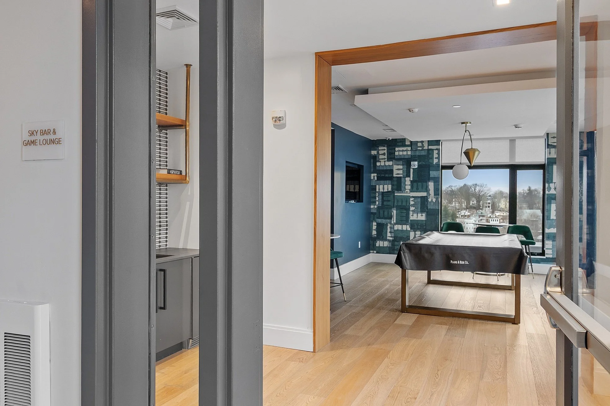 Interior of an office or lounge area with a pool table covered with a black cover, a blue accent wall with a window view, green chairs, and modern decor.