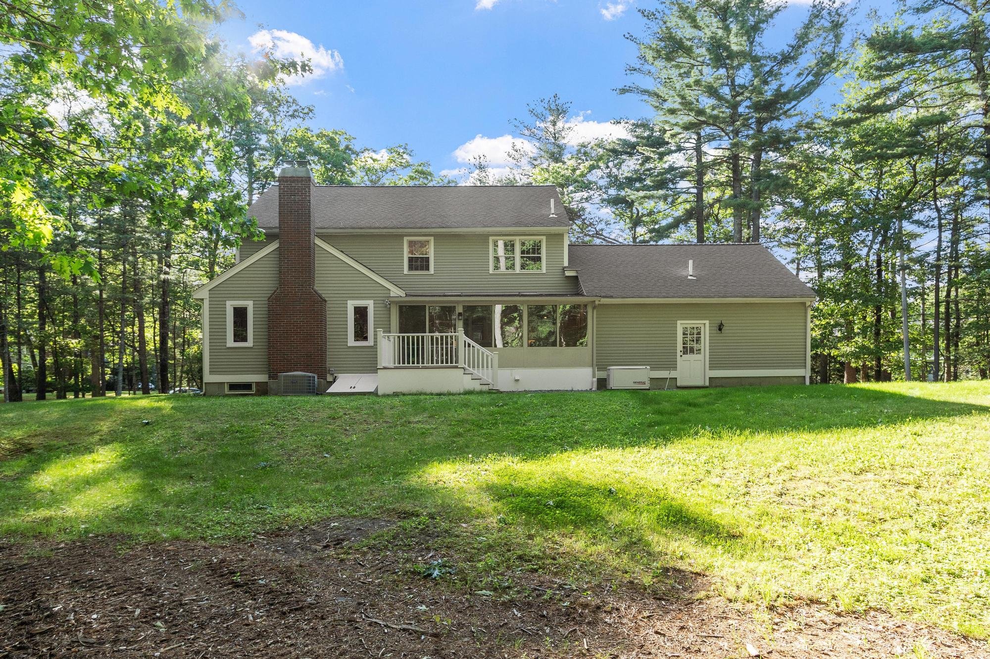 Backyard view of a two-story house with beige siding, a brick chimney, and a screened porch, surrounded by tall trees and a green lawn.