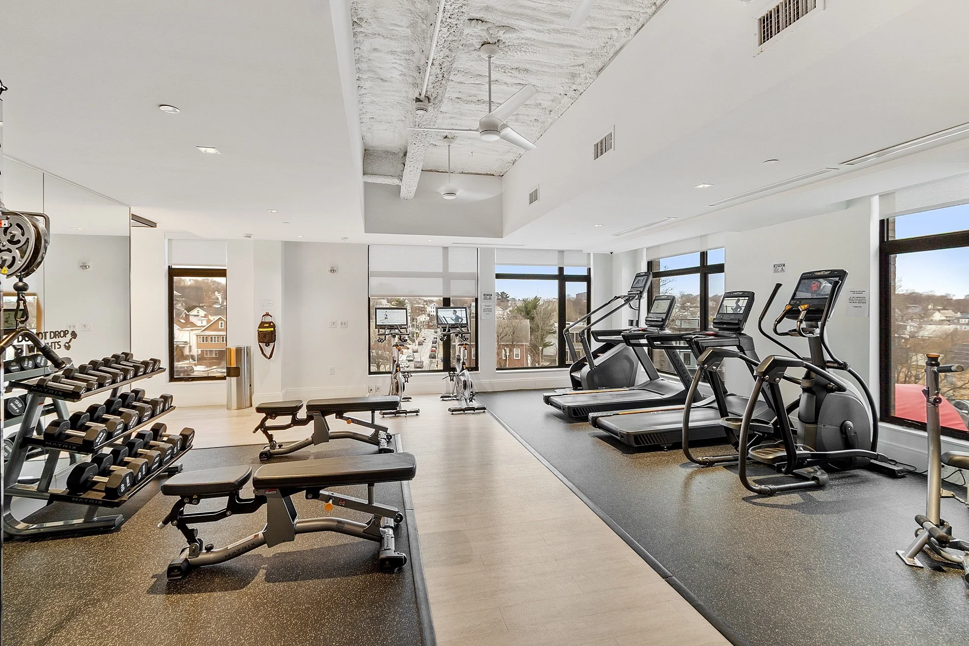 Fitness room with treadmills, stationary bikes, dumbbells, and workout benches, with large windows showing a cityview outside.