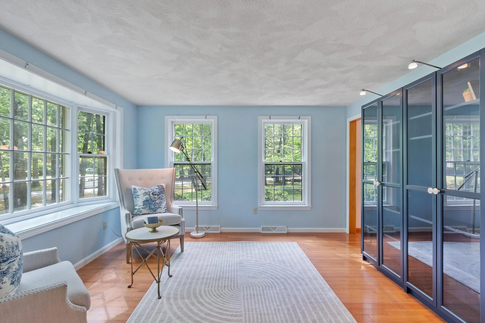 Bright living room with light blue walls, large windows showing greenery outside, a white armchair with a pillow, a small round coffee table, and a blue glass cabinet. Hardwood floors and a white area rug are visible.