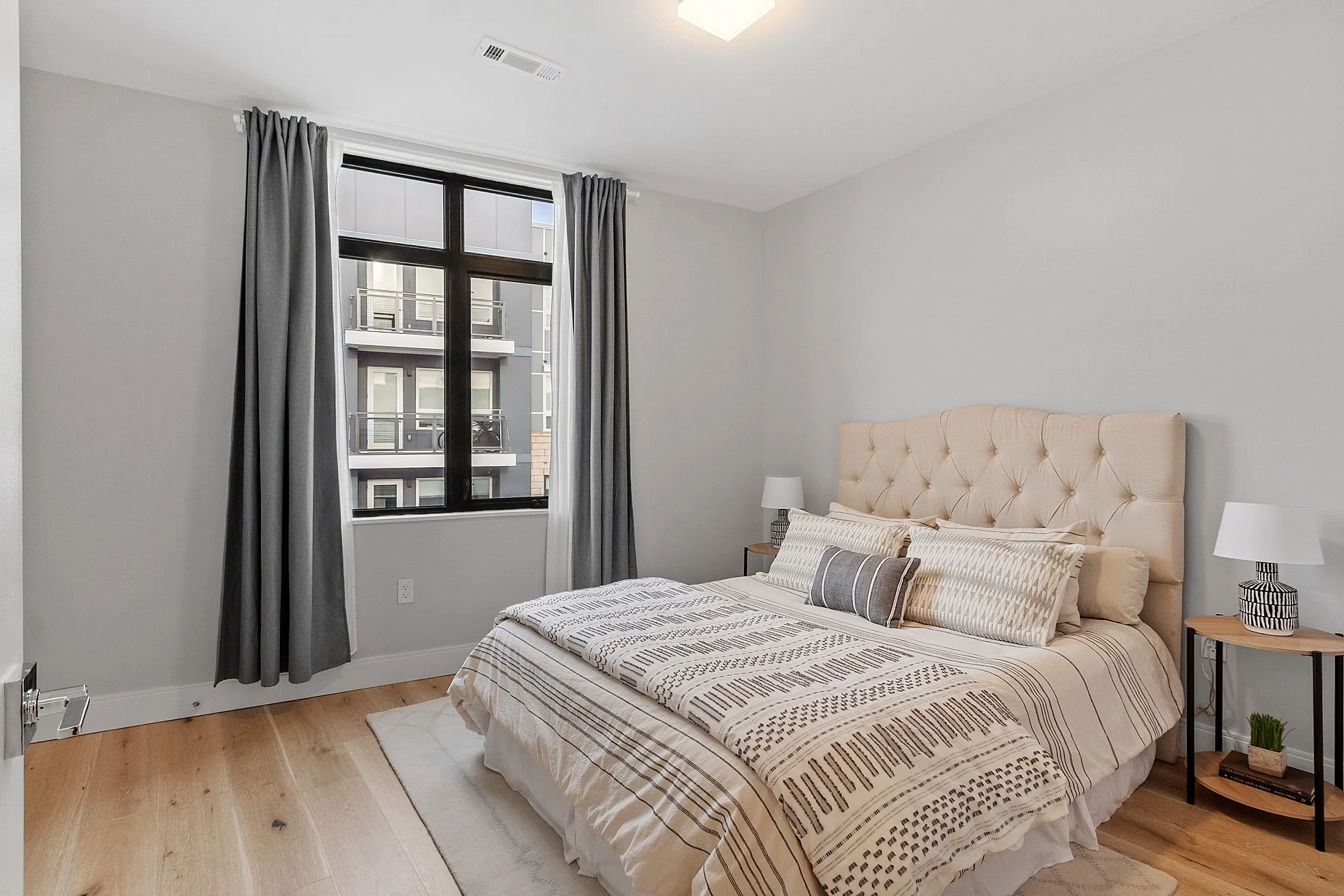 A bedroom with a large bed, beige tufted headboard, striped bedding, two nightstands with lamps, a window with gray curtains, and light-colored hardwood floors.