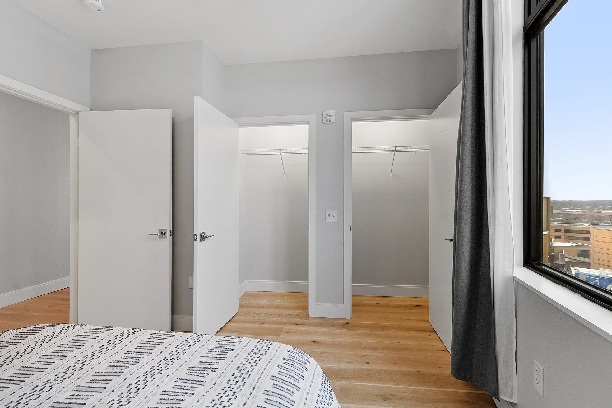 Empty bedroom with a bed in the foreground, an open closet with sliding doors, and a window with a view of buildings outside.