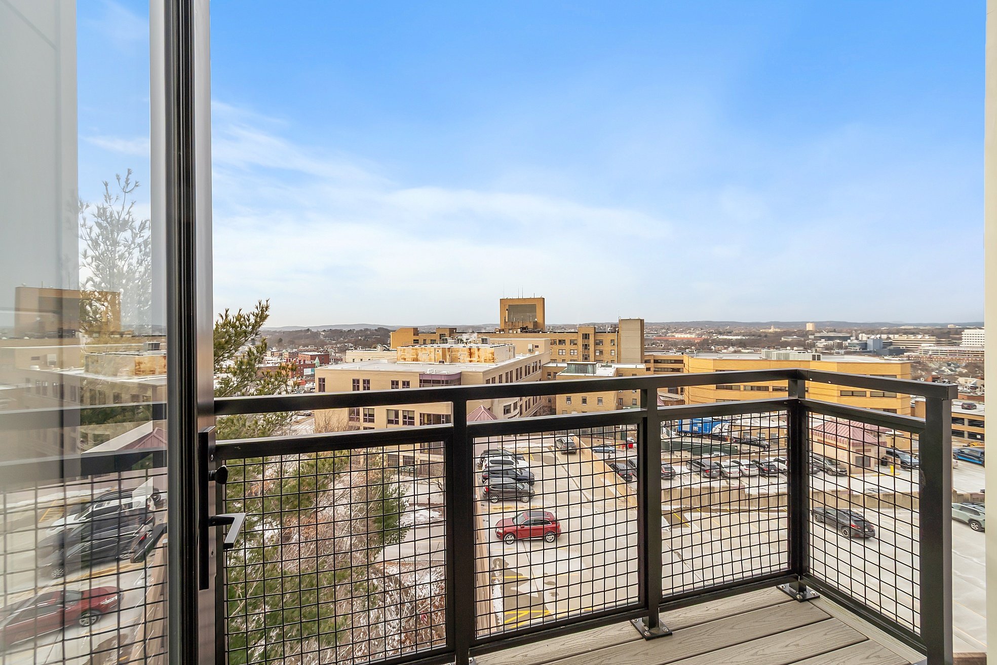 Cityscape viewed from a balcony with a metal railing, showing parking lots and beige buildings under a blue sky.