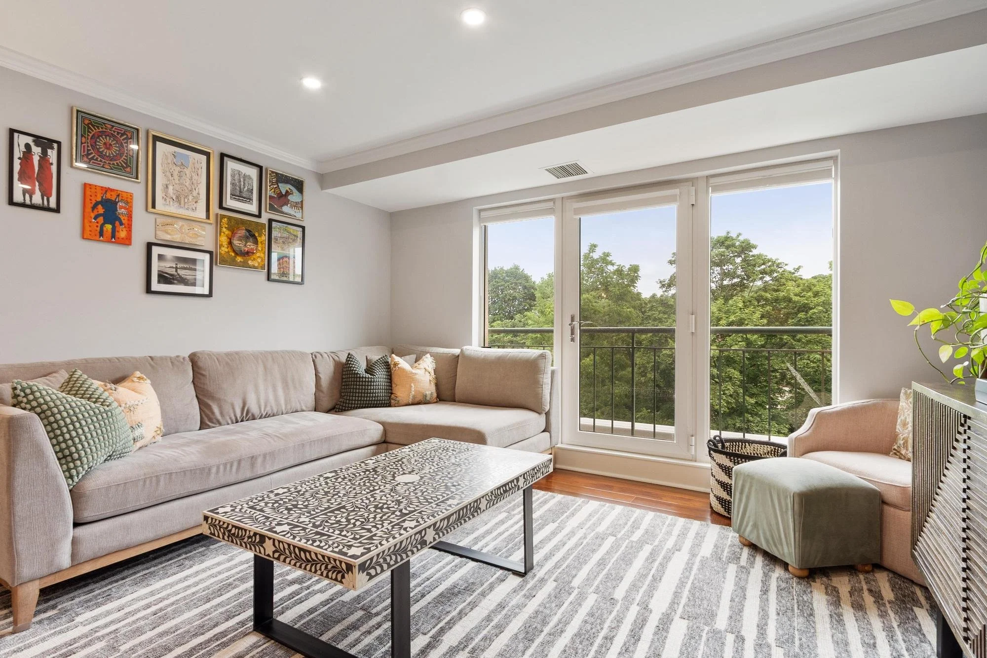 Living room with beige sofa, patterned coffee table, striped rug, wall art, large windows with greenery outside, and a plant in the corner.