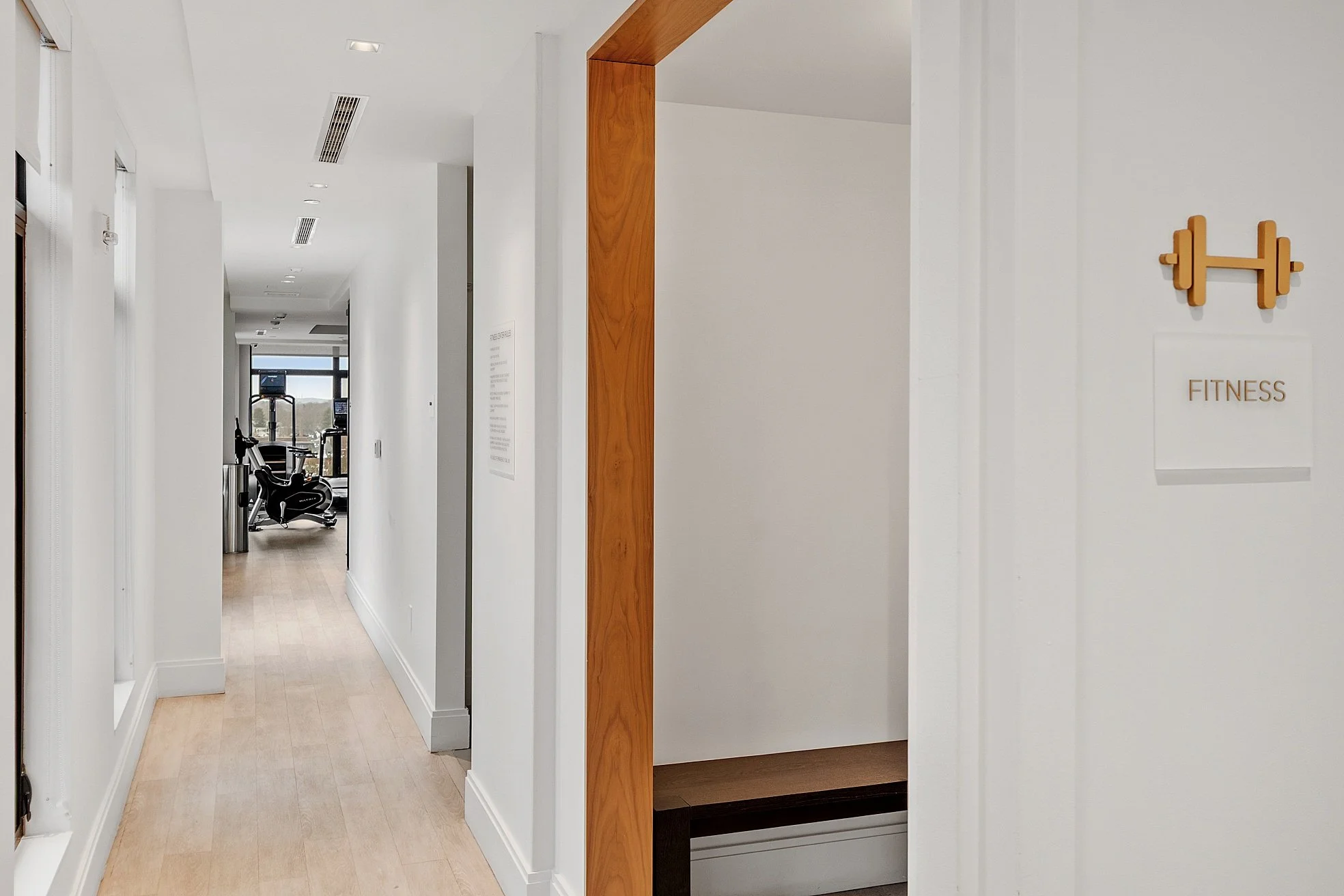 Hallway leading to a fitness area with exercise equipment, white walls, light wood floors, and a window at the end.