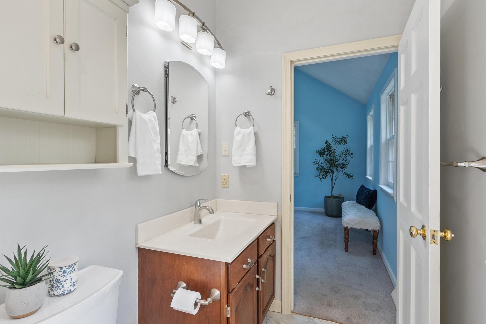 A bathroom with white walls, a wooden vanity with a sink, a mirror, and three wall-mounted light fixtures. White towels hang on towel rings, and a small potted plant and decorative containers are on the toilet tank. There is a doorway leading to a room with blue walls and a window, a potted plant, a bench, and a blue pillow.
