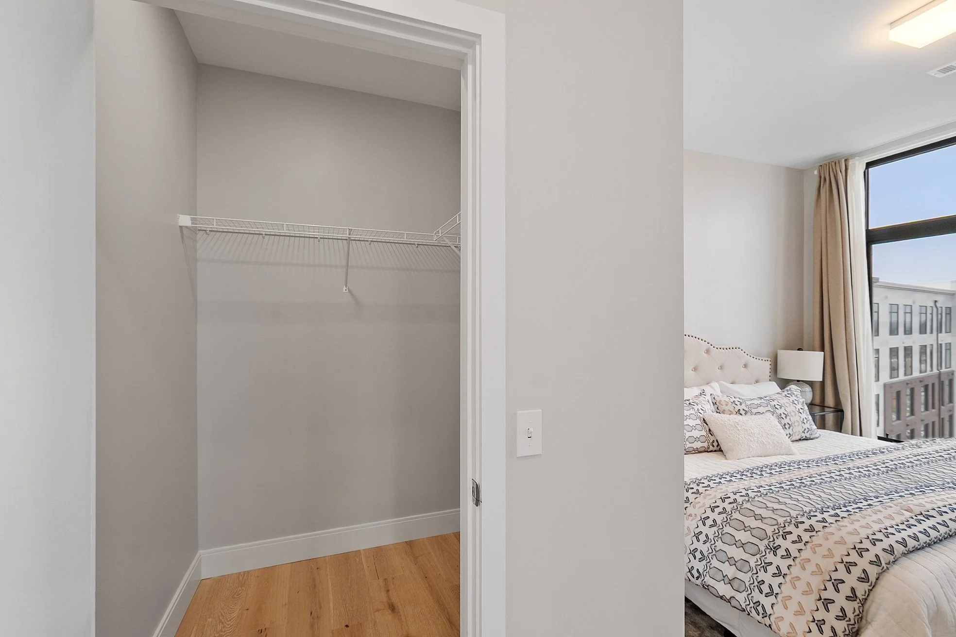 Empty closet with white wire shelf next to a bedroom with a bed, nightstand, lamp, and large window with beige curtains.