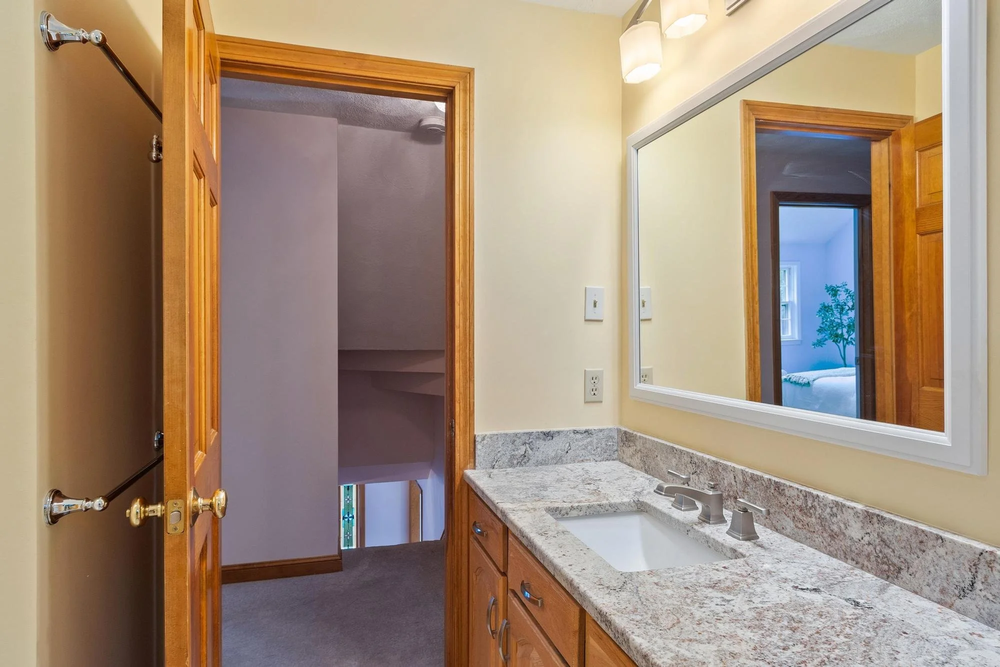 Bathroom with a granite countertop, wooden cabinets, a large mirror, and an open door revealing a closet and a bedroom with a window and a potted plant.