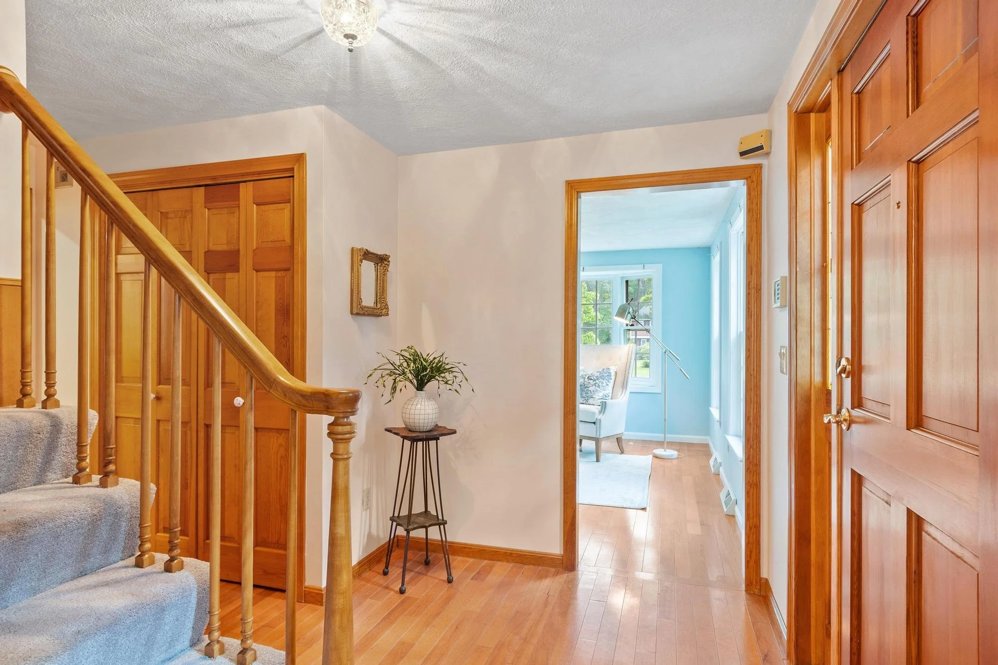 A view of a home's interior entryway showing wooden accents. There is a staircase with beige carpeted steps on the left, a small side table with a potted plant, and doorways leading to other rooms with large windows and blue walls in the background.