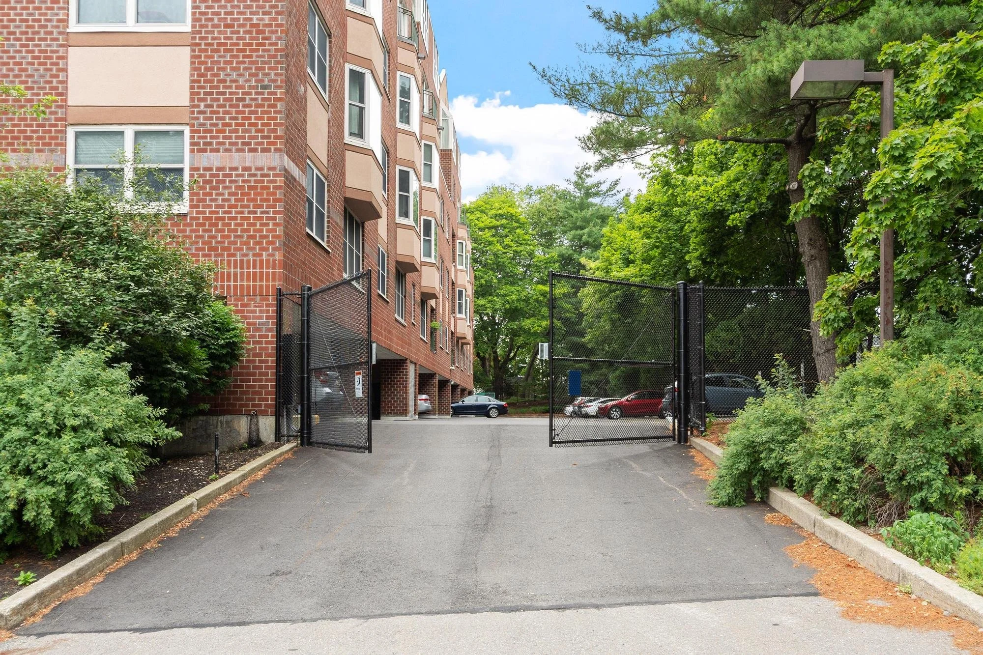 Apartment building with brick facade on the left, a parking lot behind a black fence in the center, and green trees on the right. Cars are parked in the lot and the sky is partly cloudy.