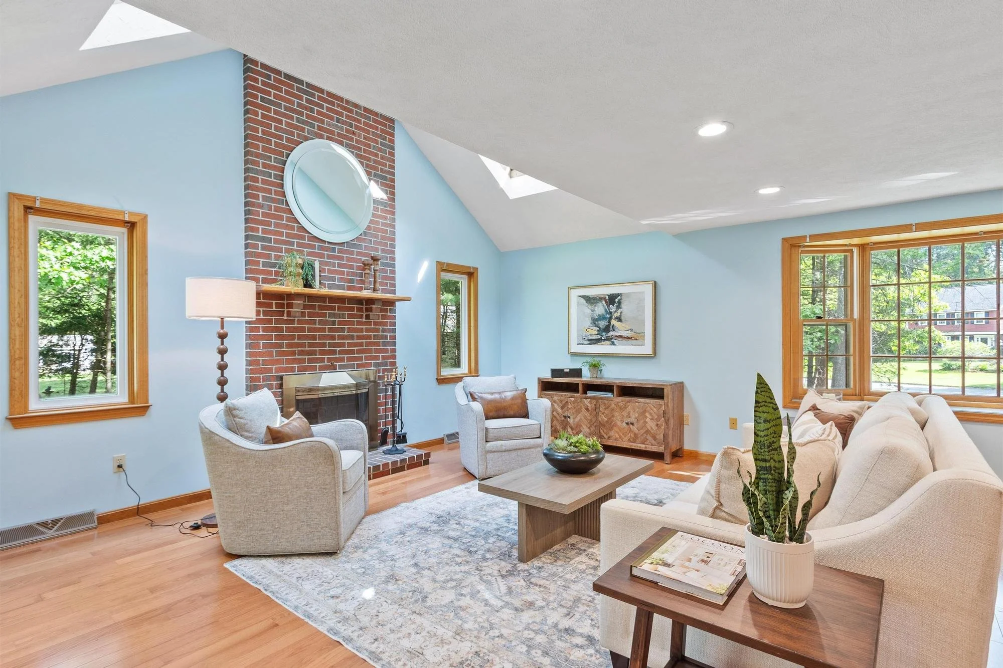 Living room with light blue walls, large windows, a brick fireplace with a round mirror, white armchairs, a wooden coffee table and a sofa, decorative plants, and artwork on the walls.