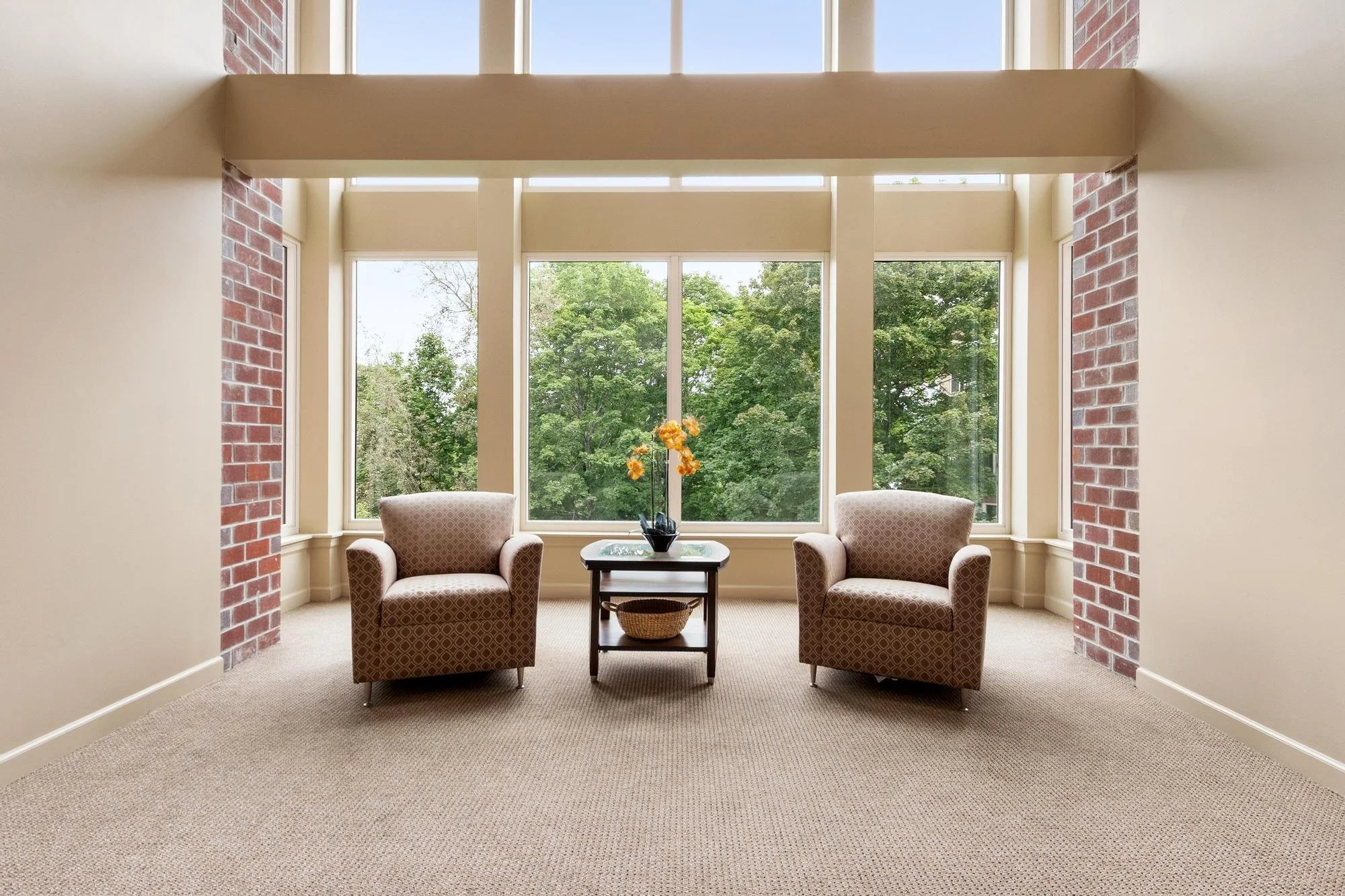 Living room with large windows overlooking green trees, two armchairs, a small table with a flower arrangement, and a carpeted floor.
