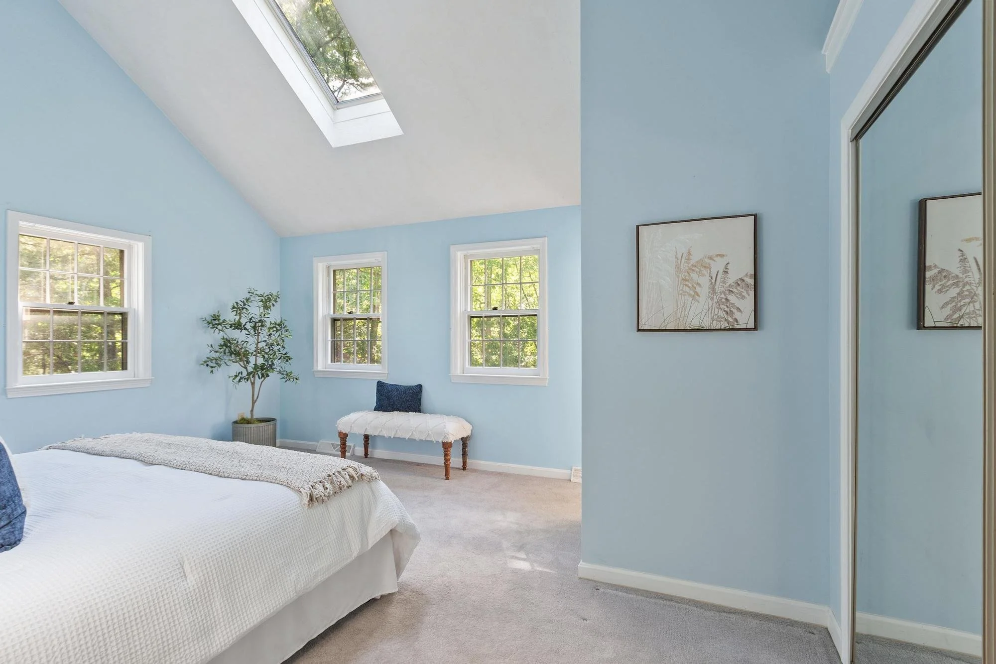 A bedroom with light blue walls, three windows, and a skylight. There is a bed with white bedding, a potted plant, a small bench with a white cushion, and framed artwork on the wall.