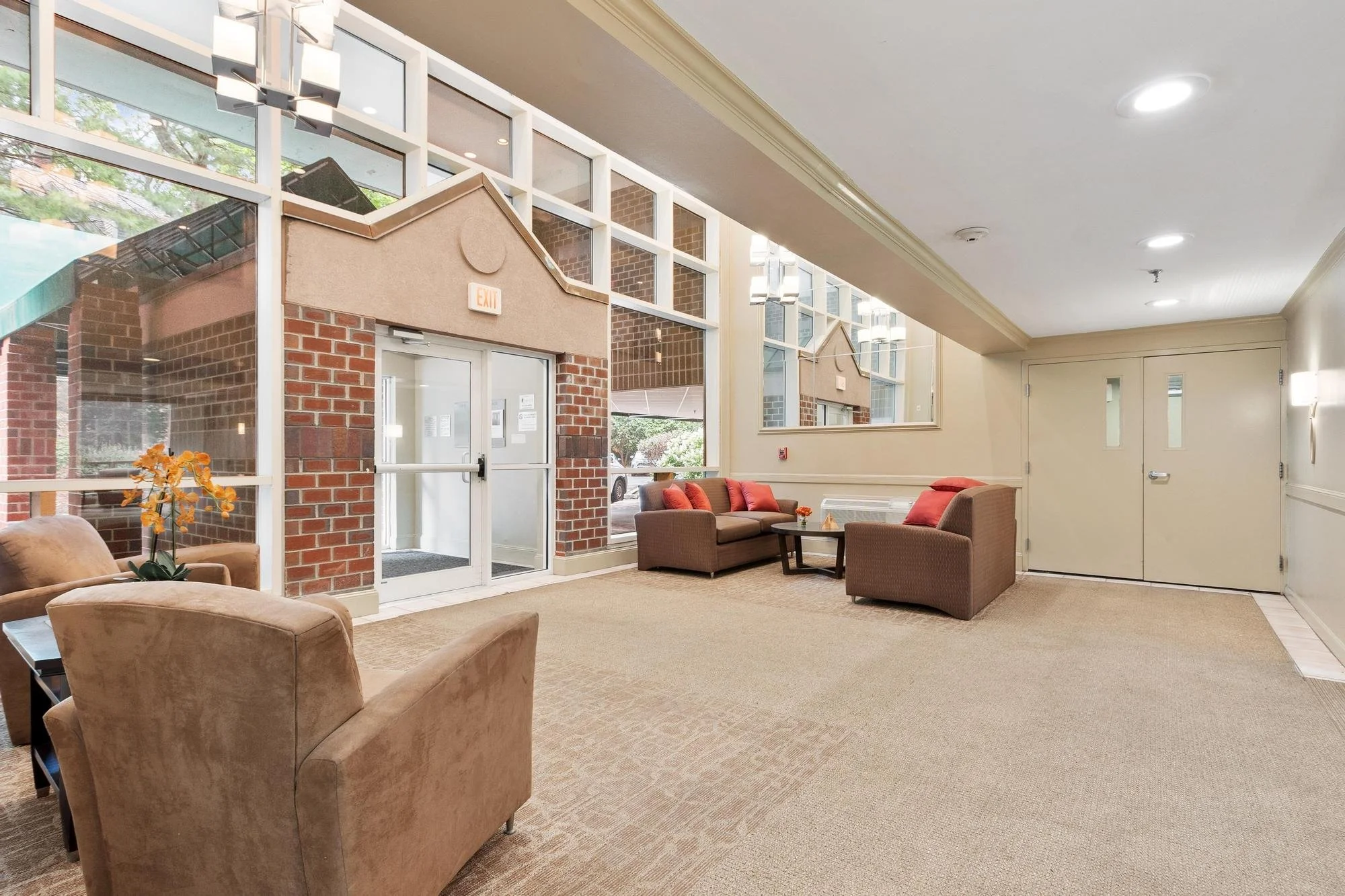 A spacious indoor lobby area with beige carpeted floor, beige chairs, and a glass wall with brick accents. There are red cushions on the chairs, a small table with a flower, and a large window with outdoor view of trees and buildings. The ceiling has recessed lighting, and there are large double doors at the back.
