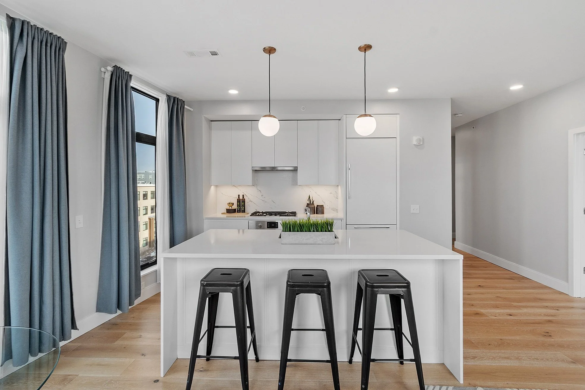 Modern kitchen with white island, three black barstools, gray curtains, light hardwood flooring, and white cabinets with marble backsplash.