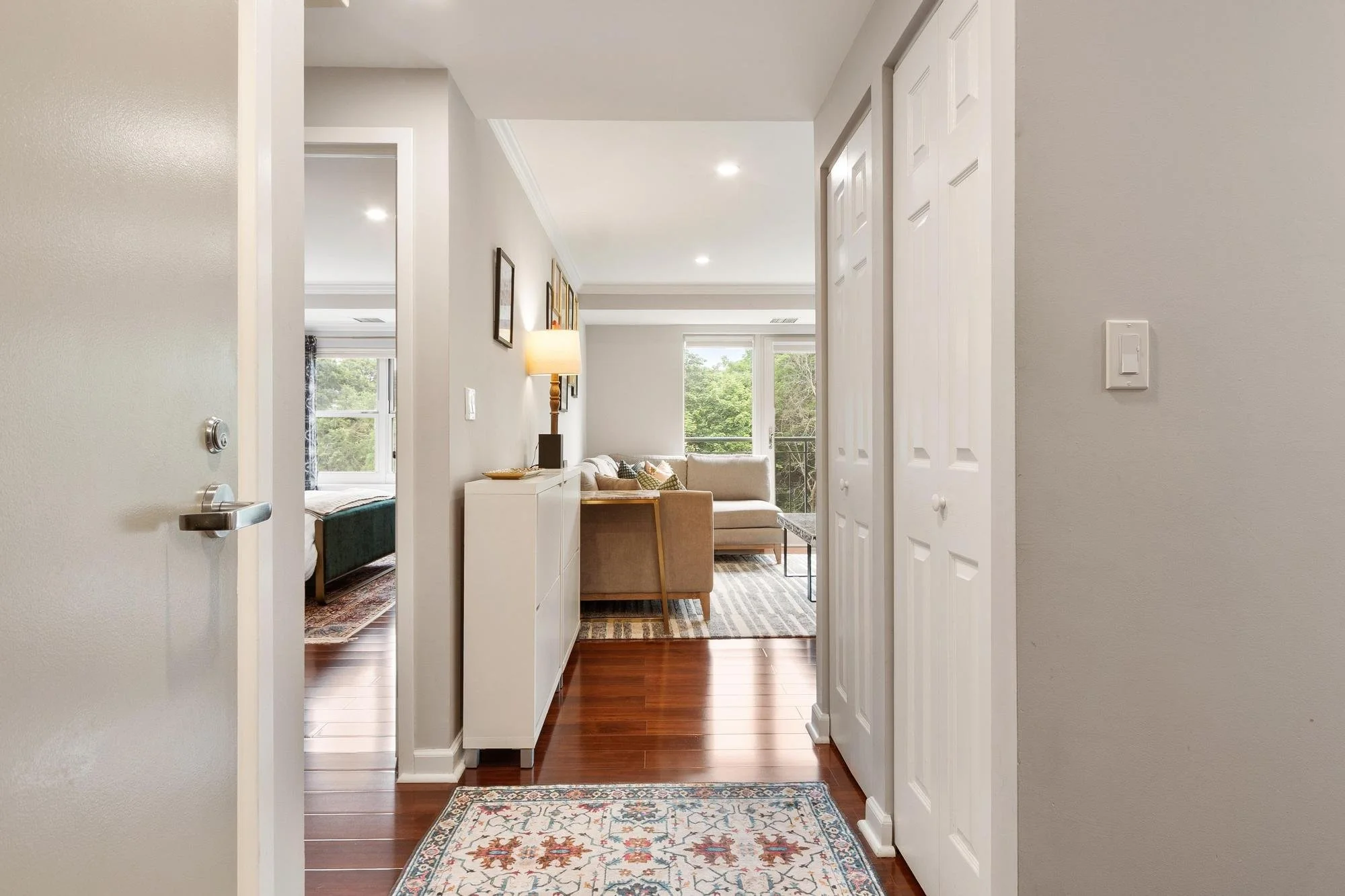 Entryway view into a living room with white furniture, a patterned rug, and large windows showing green outdoor scenery.