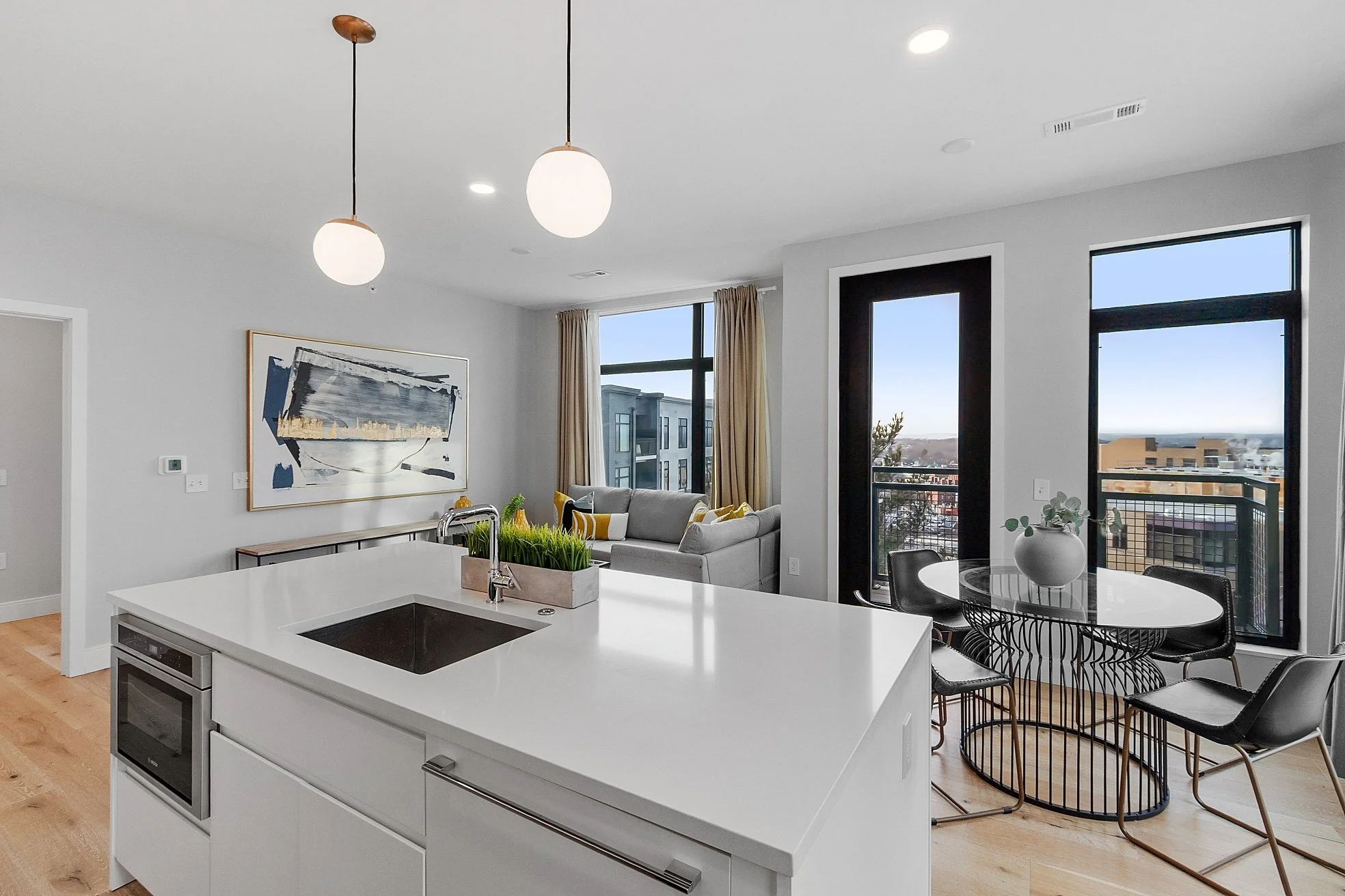 Modern open-concept kitchen and living room with white countertops, gray sofa, large windows, and a small dining table with four black chairs.