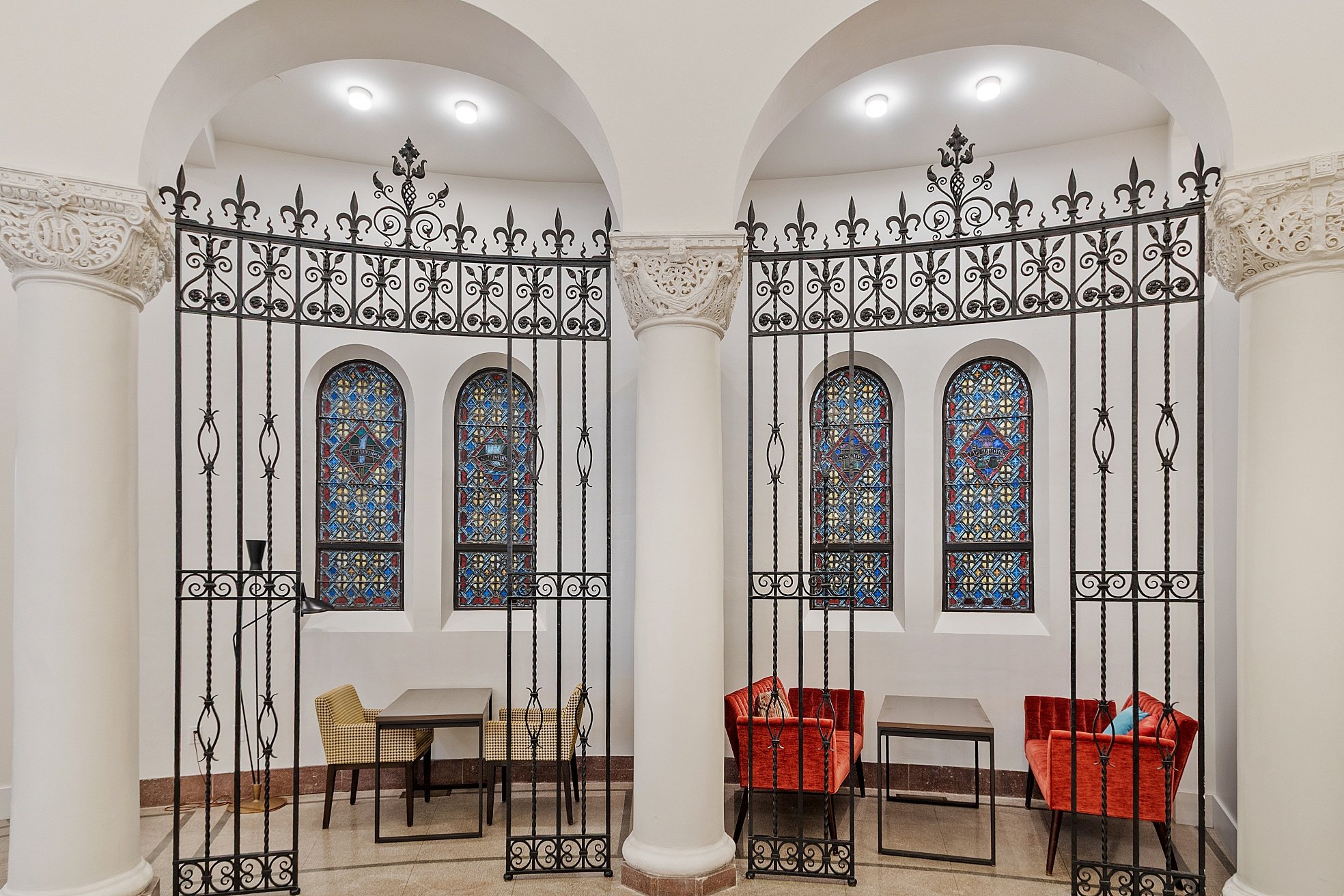 Interior of a room divided by ornate black iron gates, with stained glass windows, white columns, and seating areas with chairs and tables.