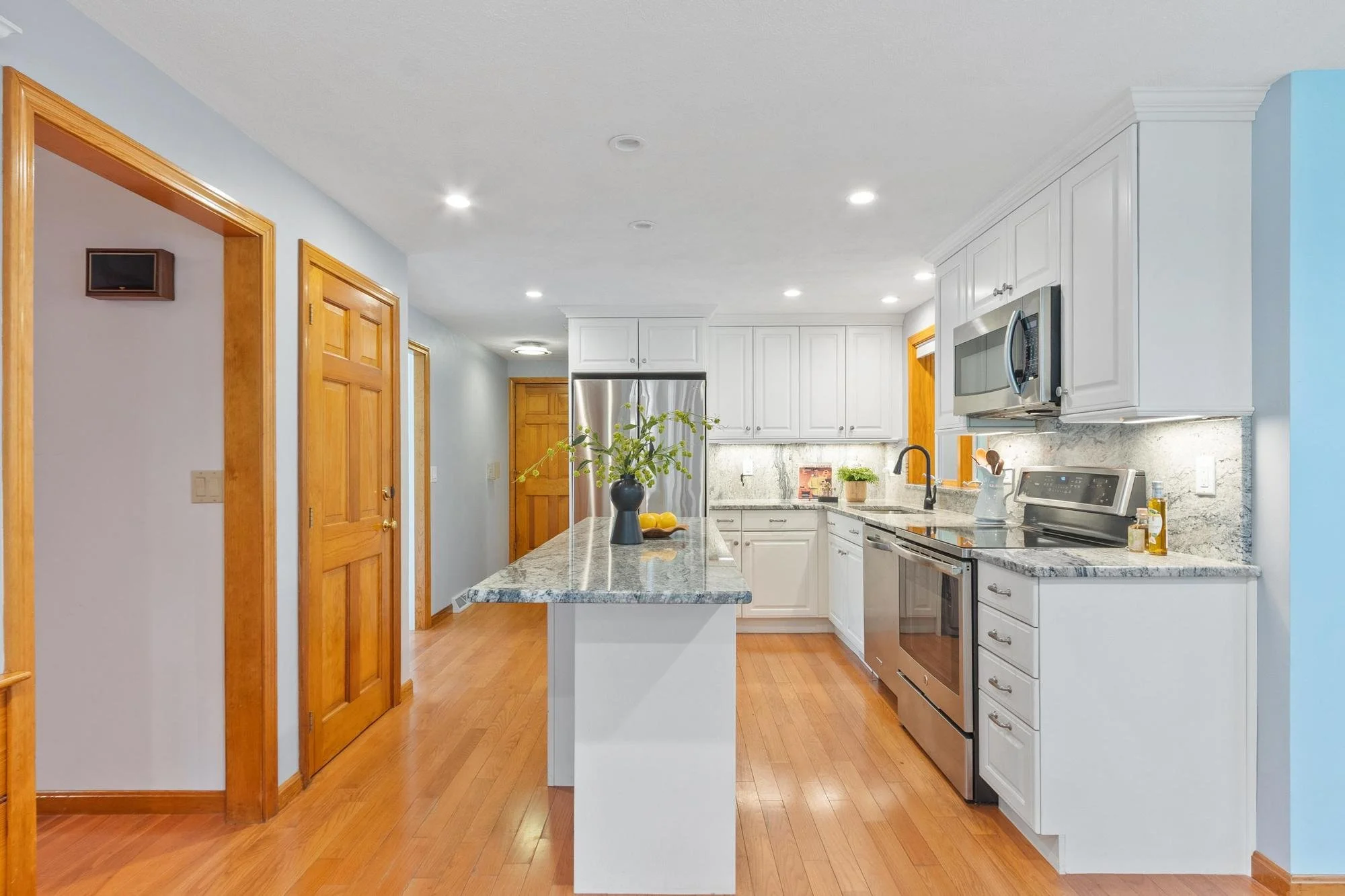 Modern kitchen with white cabinets, stainless steel appliances, marble countertops, and hardwood floors. A kitchen island with a vase of flowers and lemons, and a blue wall on the right.