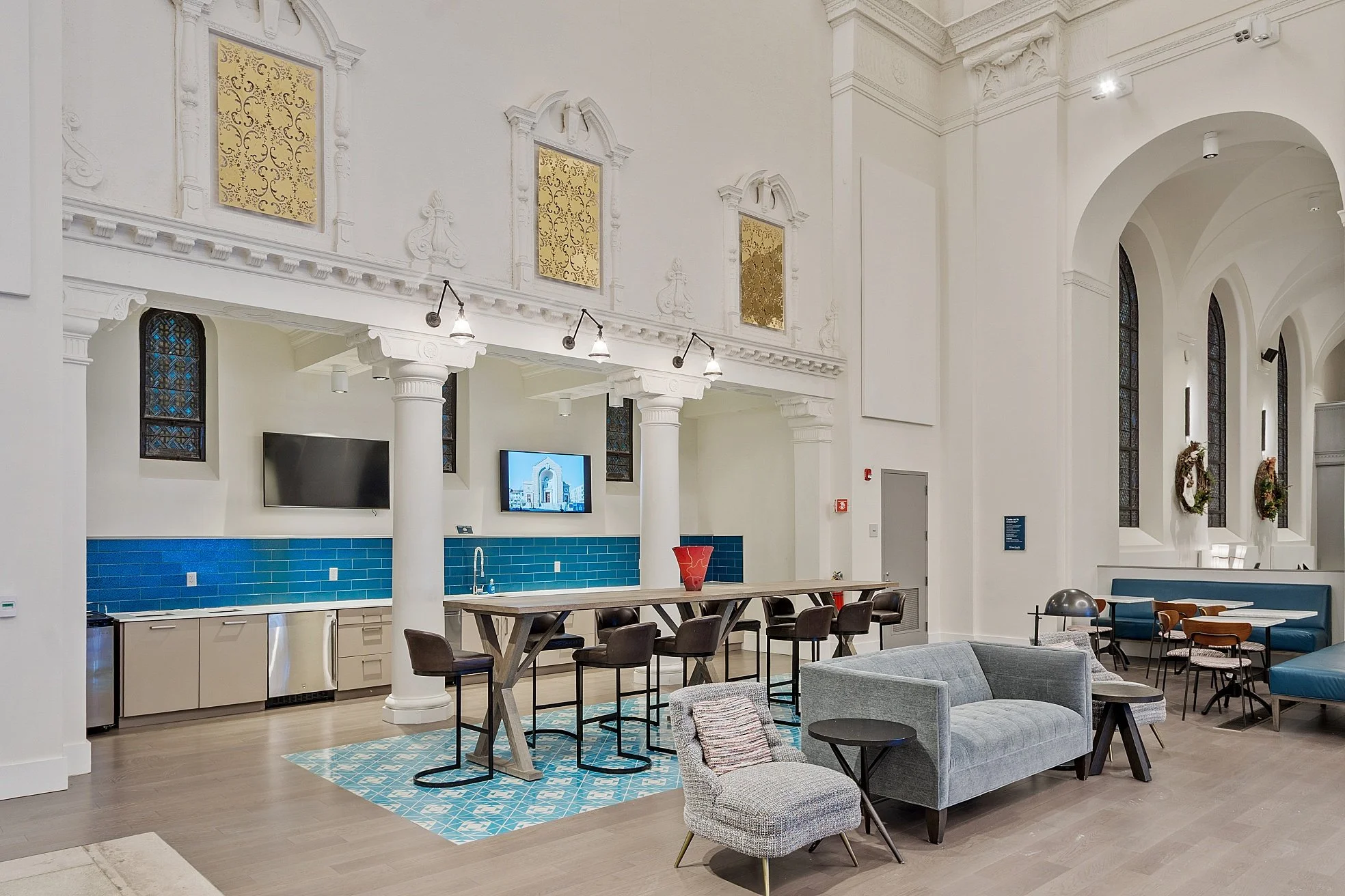 Indoor lounge area with high ceilings, stained glass windows, and white walls. Features a kitchenette with blue backsplash, television screens, a high table with stools, and comfortable seating including gray and blue couches, chairs, and tables. Decorated with wreaths and a patterned rug.