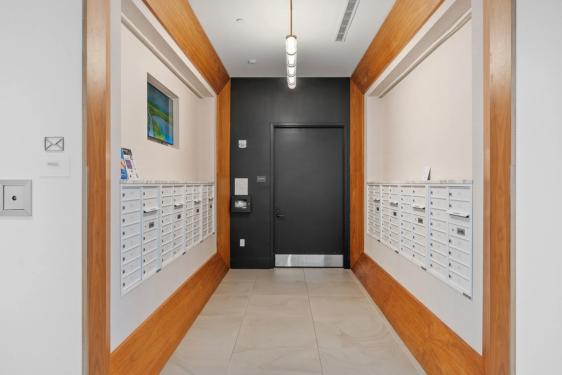 Lobby with mailboxes on both sides, a black door in the center, a wall-mounted TV on the left, and a light fixture hanging from the ceiling.