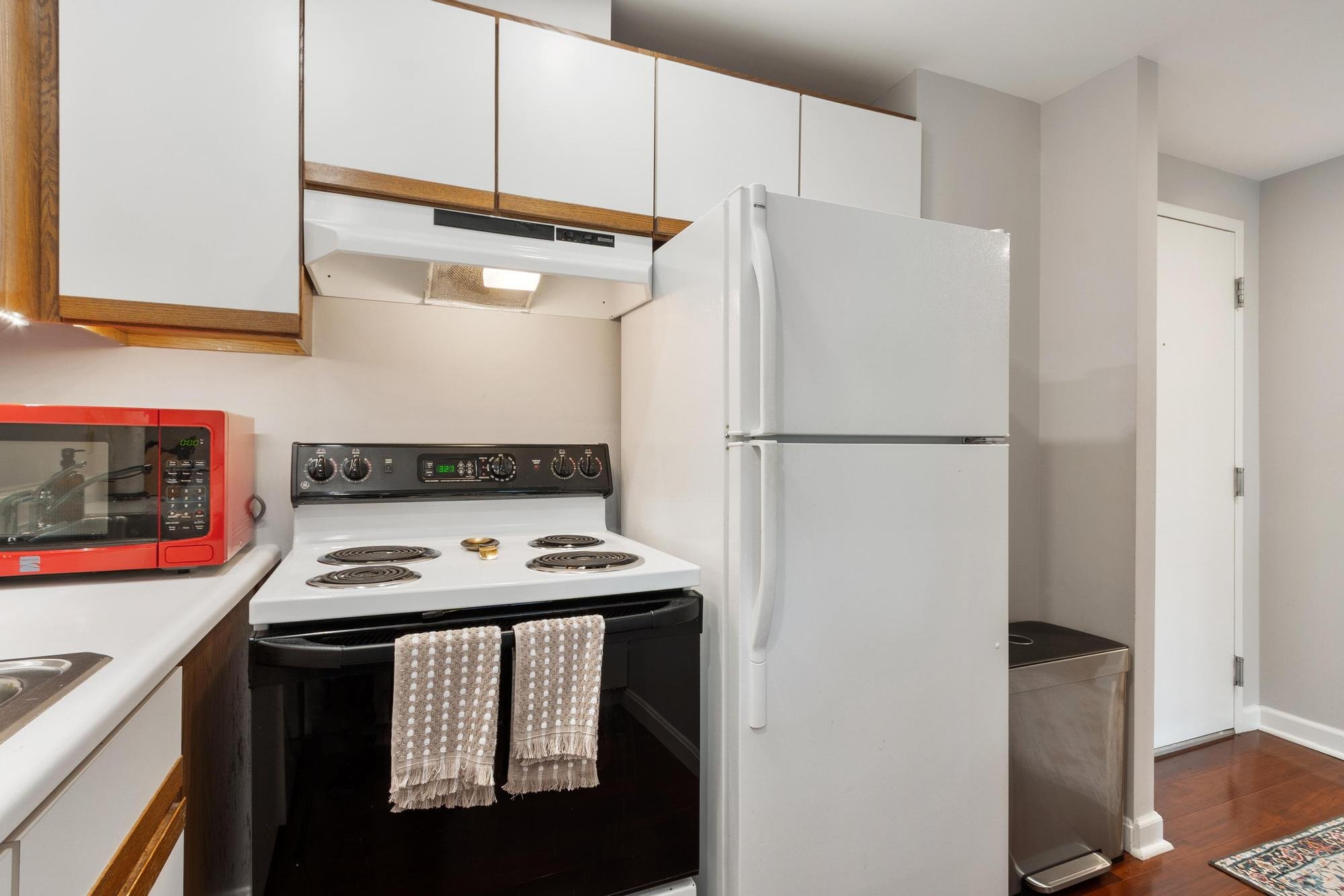 A kitchen with white cabinets and appliances, including a white refrigerator and a stove. There is a red microwave on the counter, and towels hanging on the oven handle. Wooden trim accents the cabinetry, and a small rug is on the floor near the door.