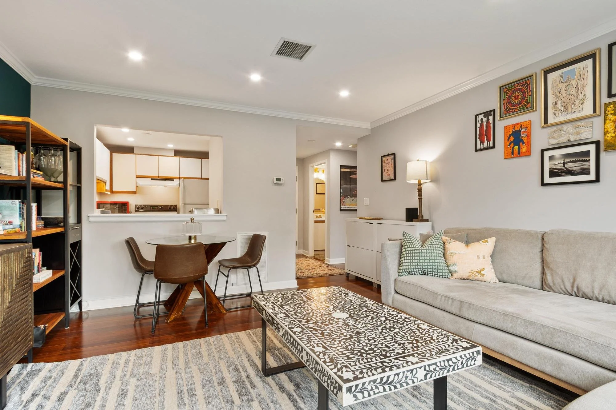 Interior view of a living room with a gray sofa, patterned coffee table, gallery wall, lap lamp, and a view into an open kitchen and dining area.
