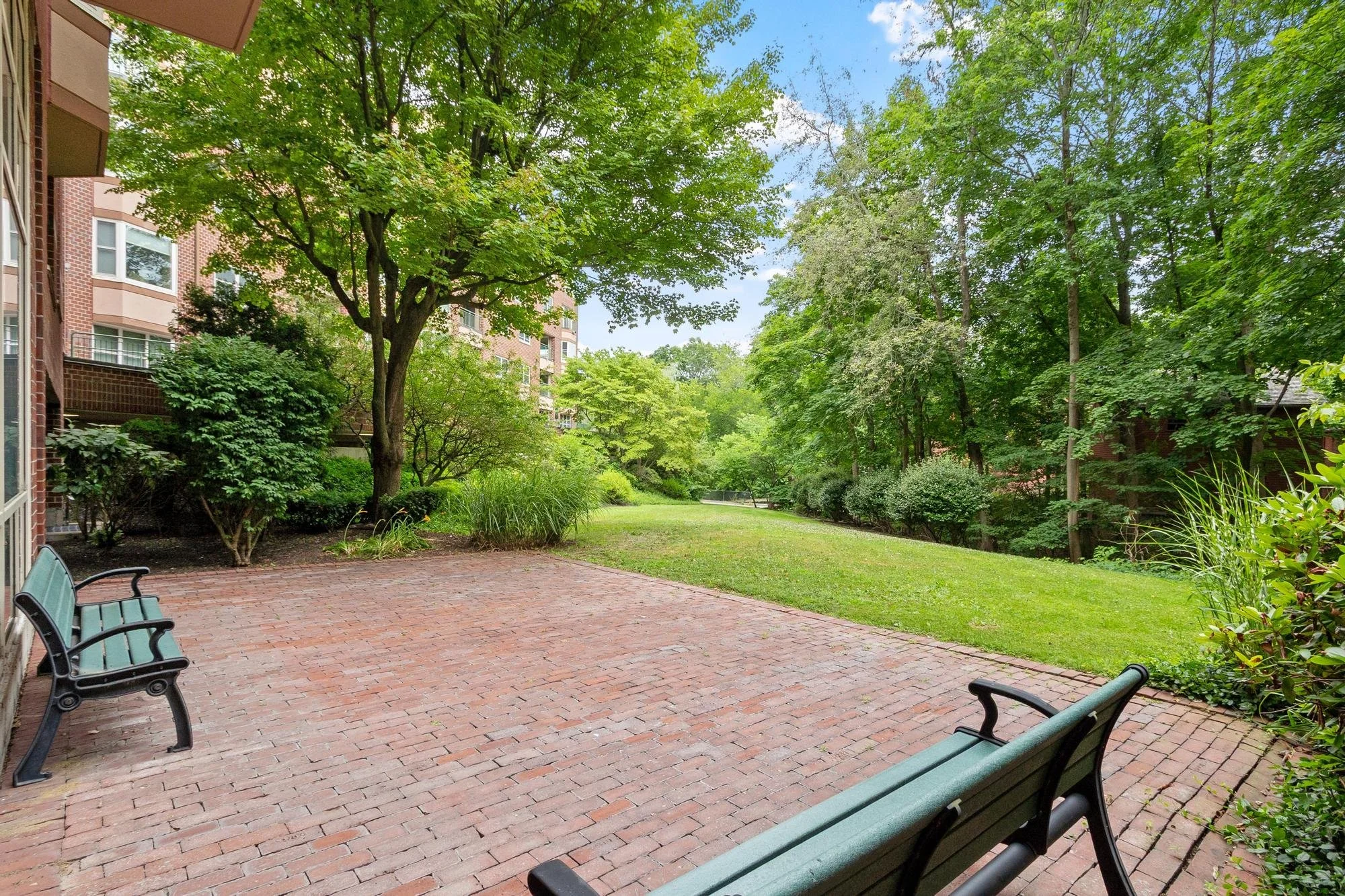 A serene outdoor courtyard with a brick patio surrounded by lush green trees and bushes. Several green benches are placed along the patio, and an apartment building with multiple windows and balconies is visible on the left side.