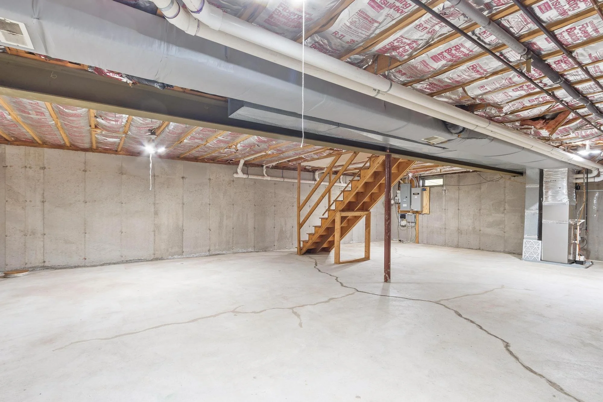 Unfinished basement with exposed ceiling insulation, ductwork, and electrical panel, featuring a wooden staircase.