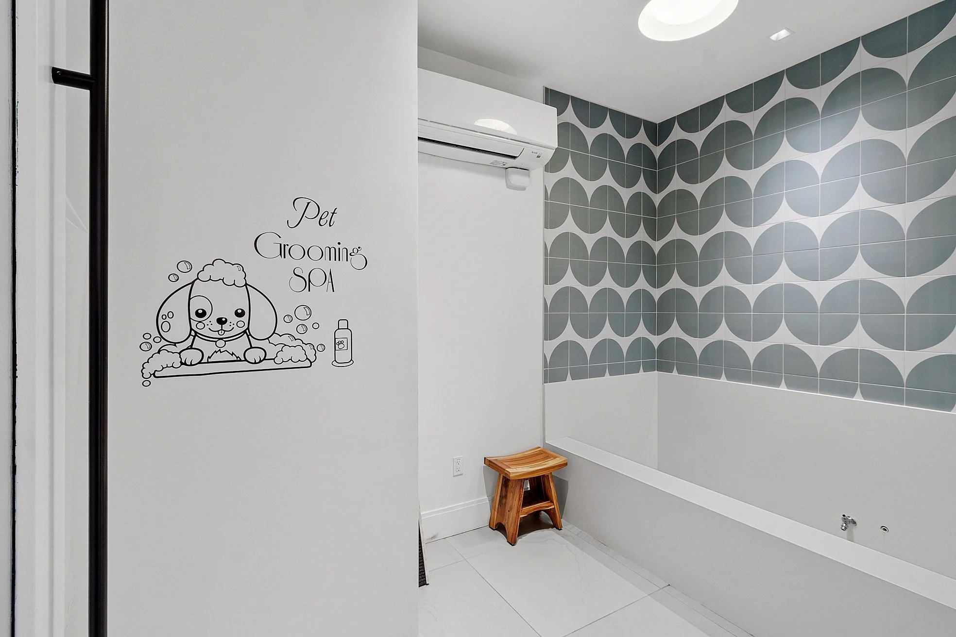 Interior of a pet grooming spa with a wall decal featuring a cartoon dog in a bath, and text reading 'Pet Grooming SPA'. The room has geometric-patterned wall tiles in shades of gray, a small wooden bench, a white wall-mounted air conditioner, and a white floor.