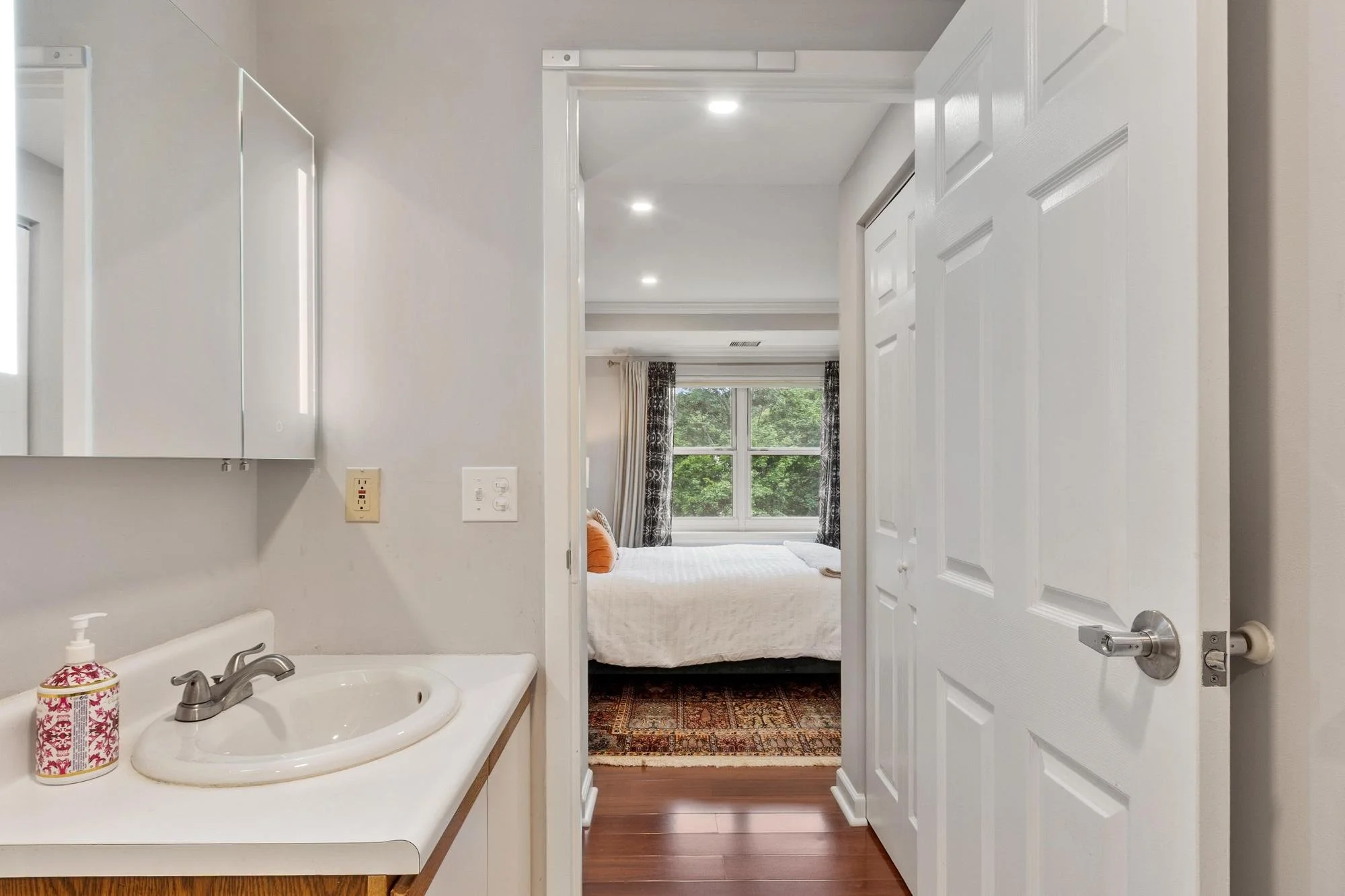 Bathroom with a sink and a soap dispenser on the counter, view into a bedroom through an open door, featuring a bed with orange pillows, a window with curtains, and hardwood floors.