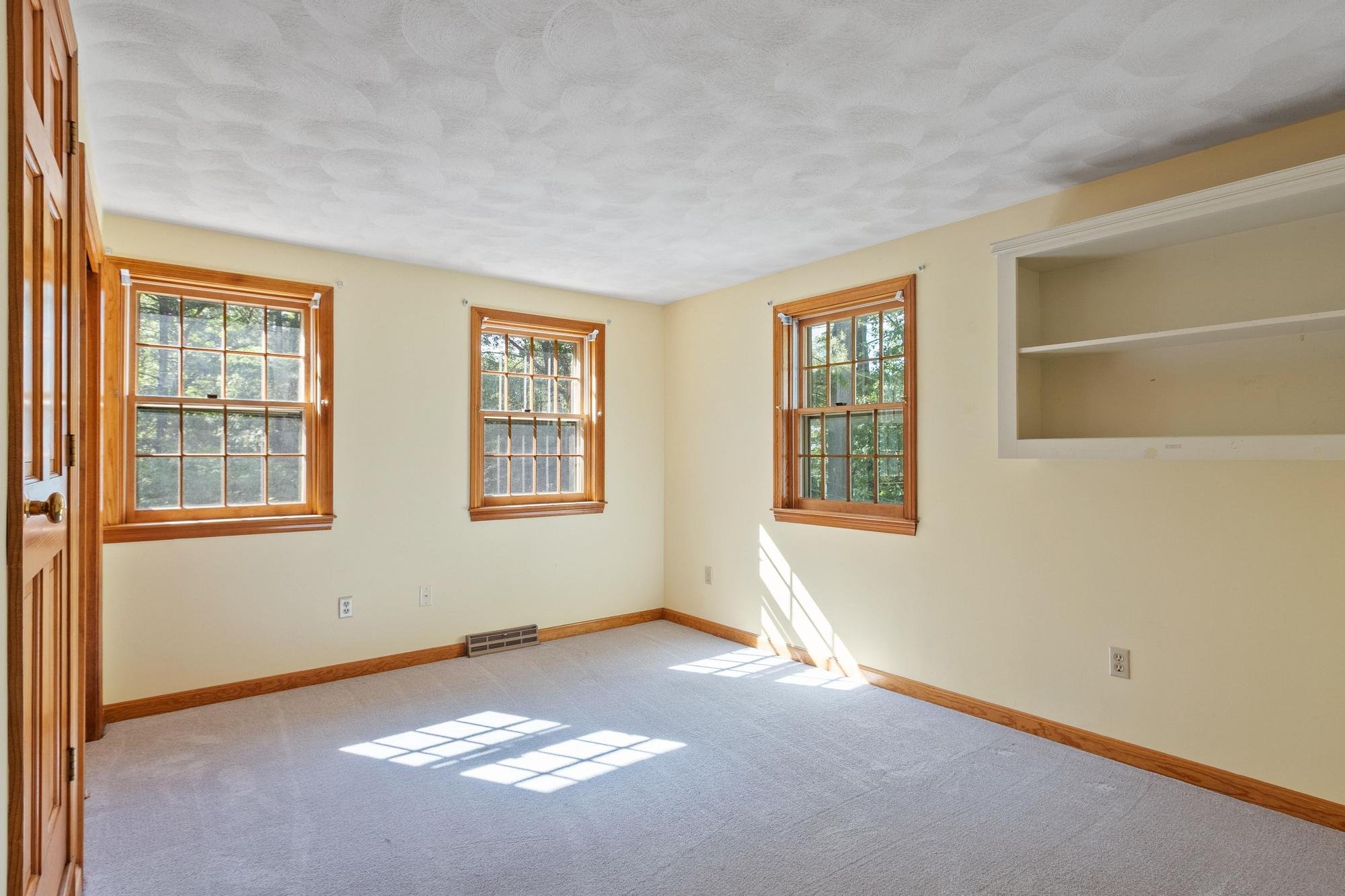 Empty room with three windows allowing natural sunlight, beige walls, wooden trim, and a white ceiling with a textured finish.