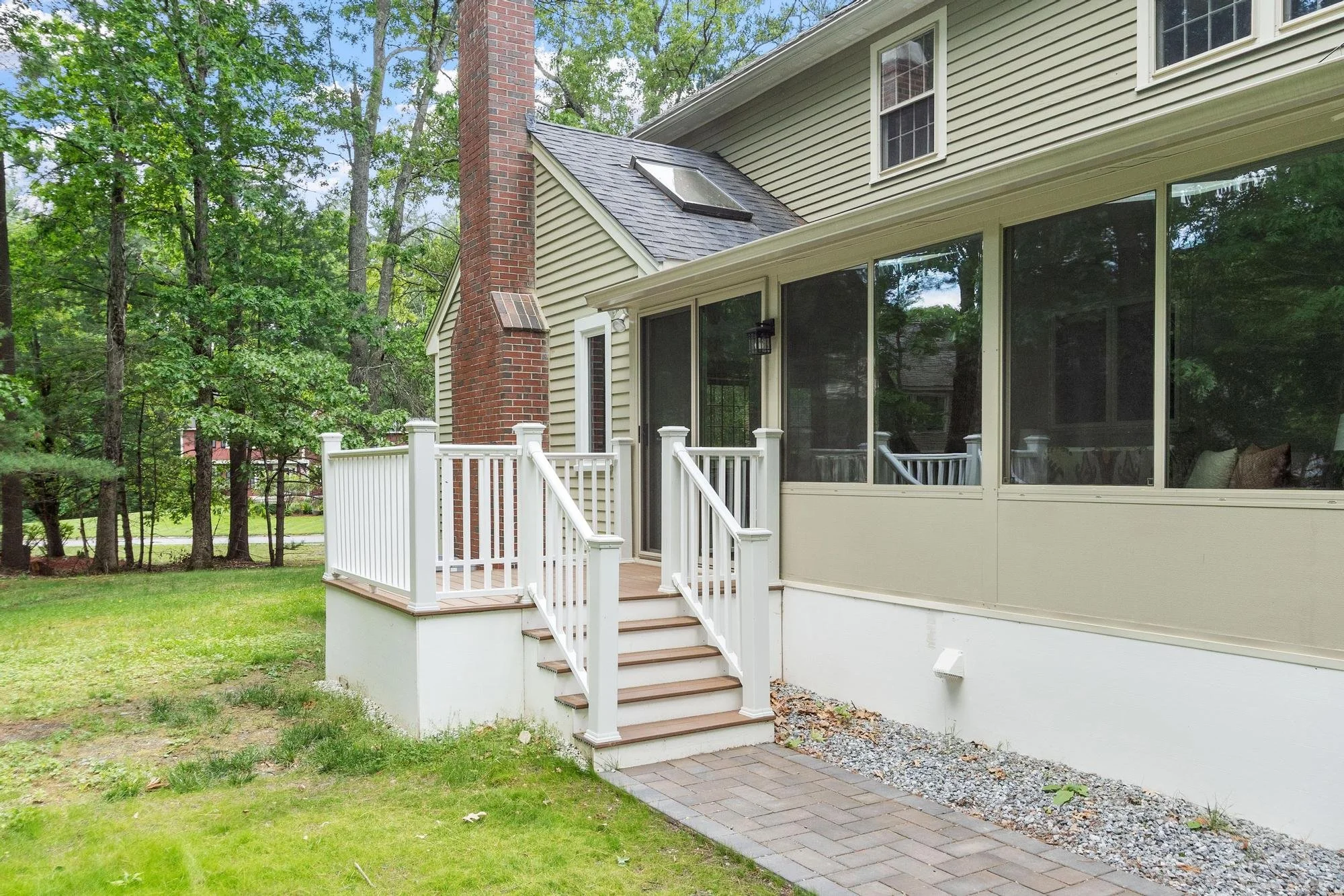 Backyard of a house with a small porch, white railing, stairs, large screened-in porch, and a brick chimney surrounded by trees and green grass.